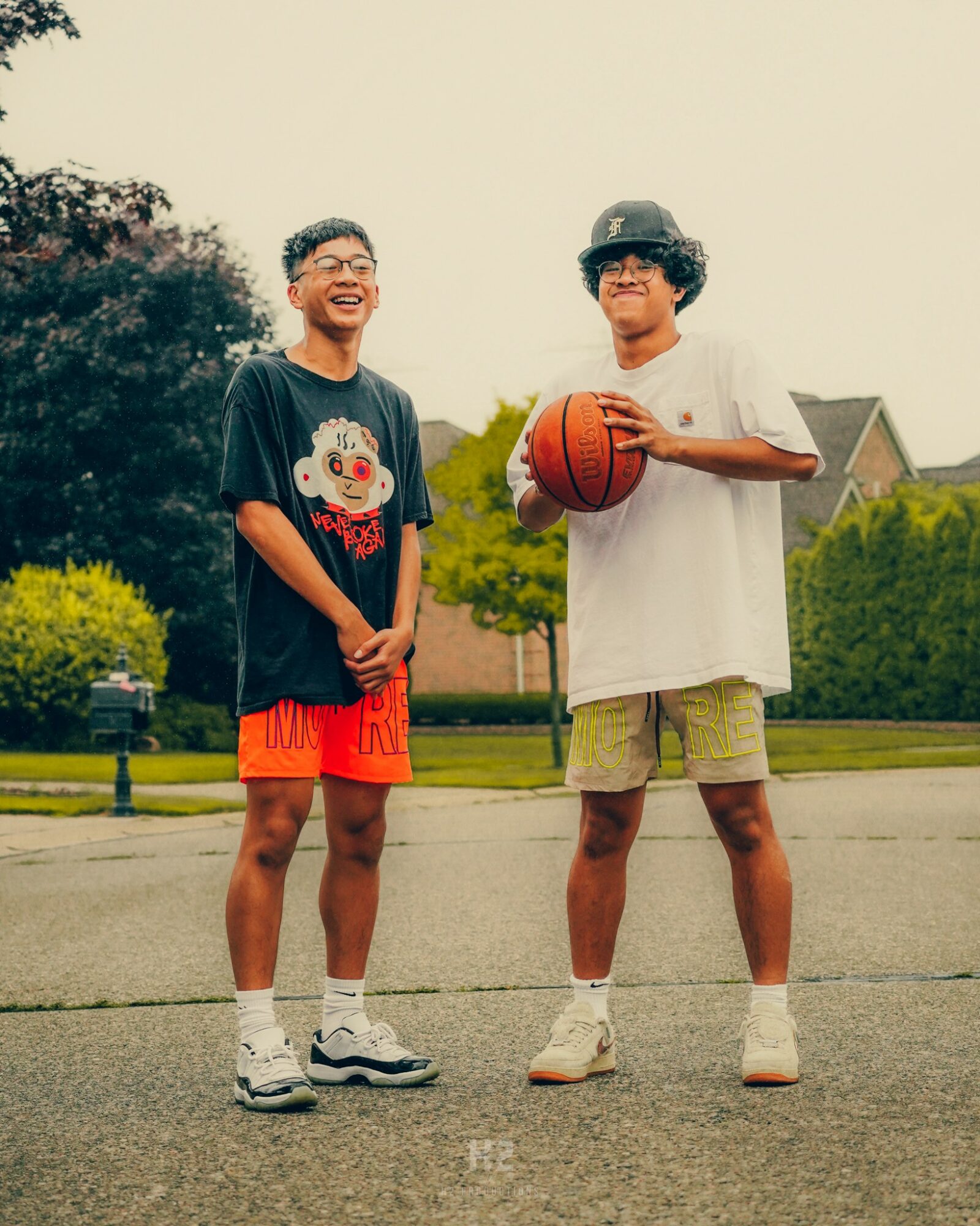 Two boys standing outdoors, one holding a basketball, smiling, with trees and houses in the background.