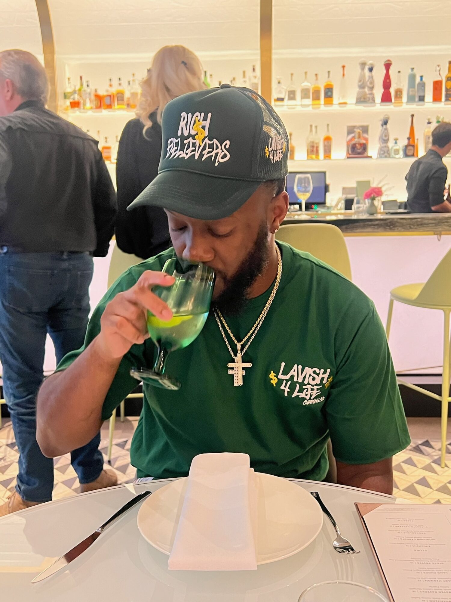 Man with a beard wearing a black cap and green shirt, drinking a green beverage in a restaurant.