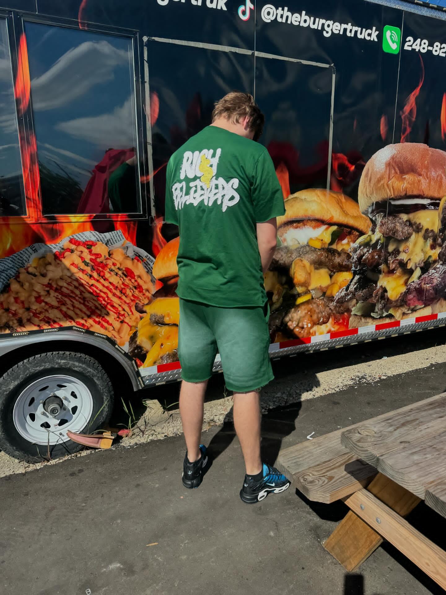 Person standing with back to camera in front of a burger-themed food truck, wearing a green shirt and shorts.