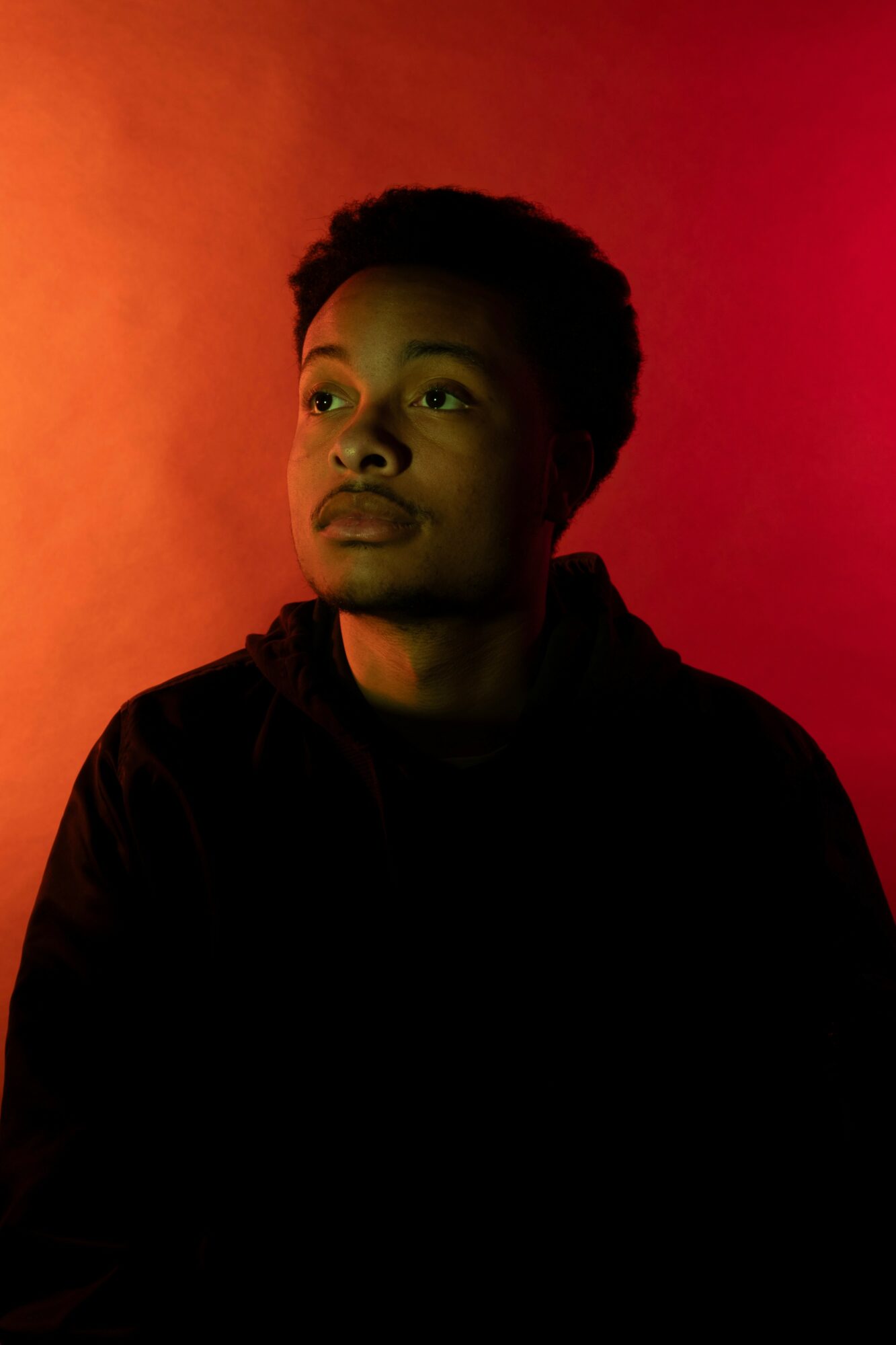 Young person with short hair looking to the side, illuminated by warm red and orange lighting, wearing a dark hoodie.