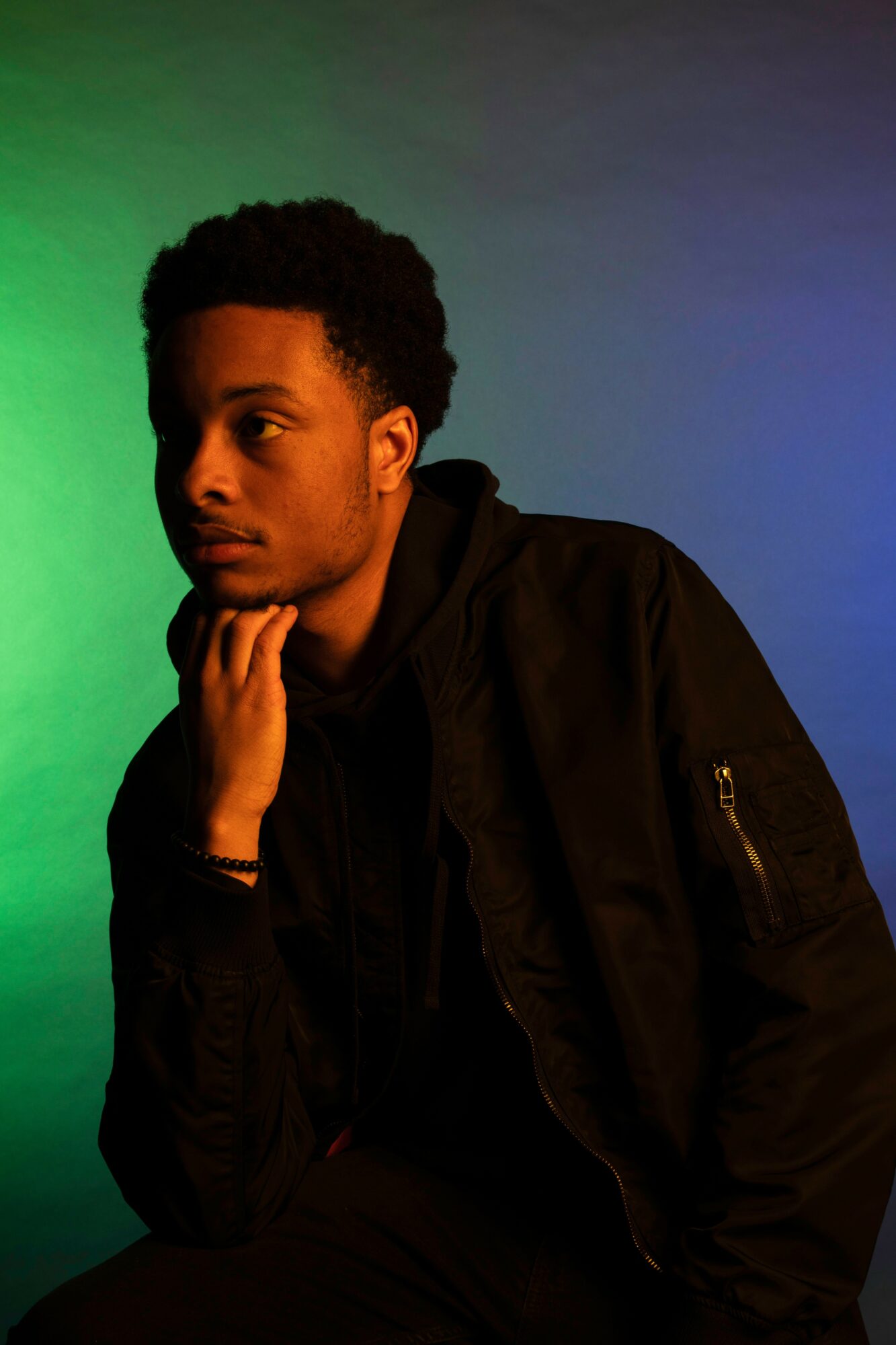 Young man with curly hair, wearing a black jacket, resting his chin on his hand, against a gradient background.