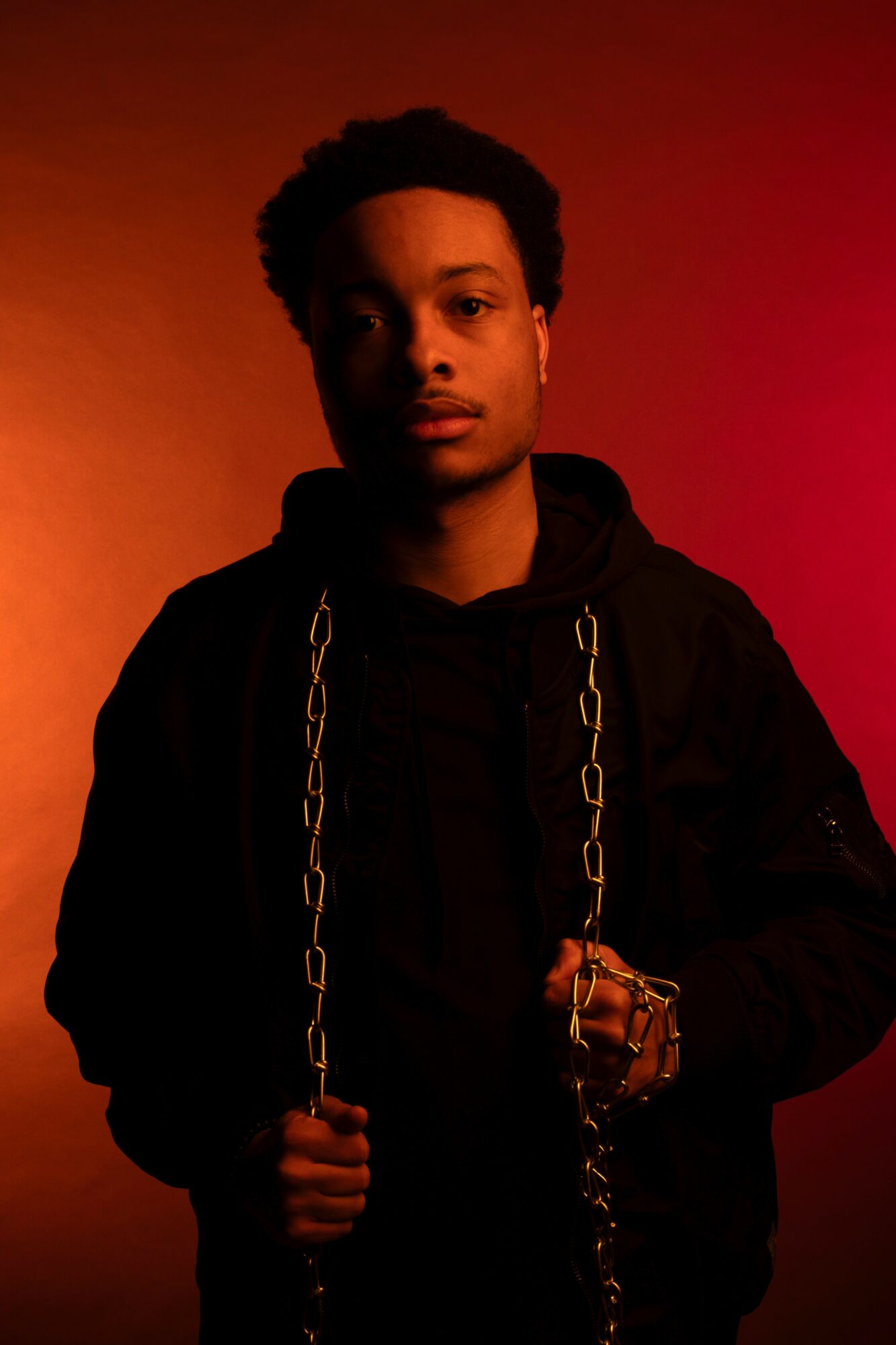 Young man with short curly hair wearing a black hoodie and jacket, holding a chain, against a red and orange background.