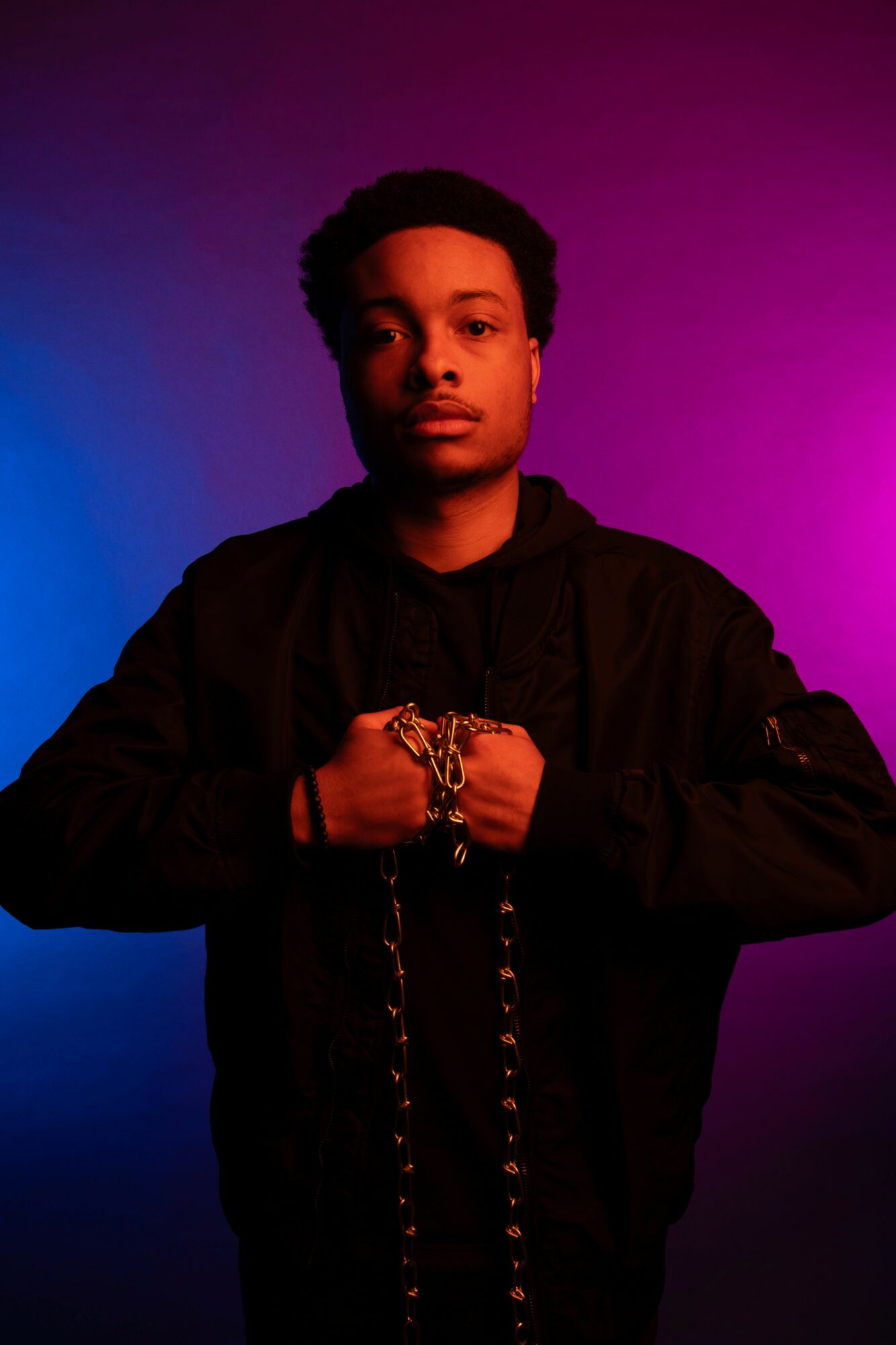 Young man with short hair wearing a black jacket, holding chains, against a colorful gradient background.