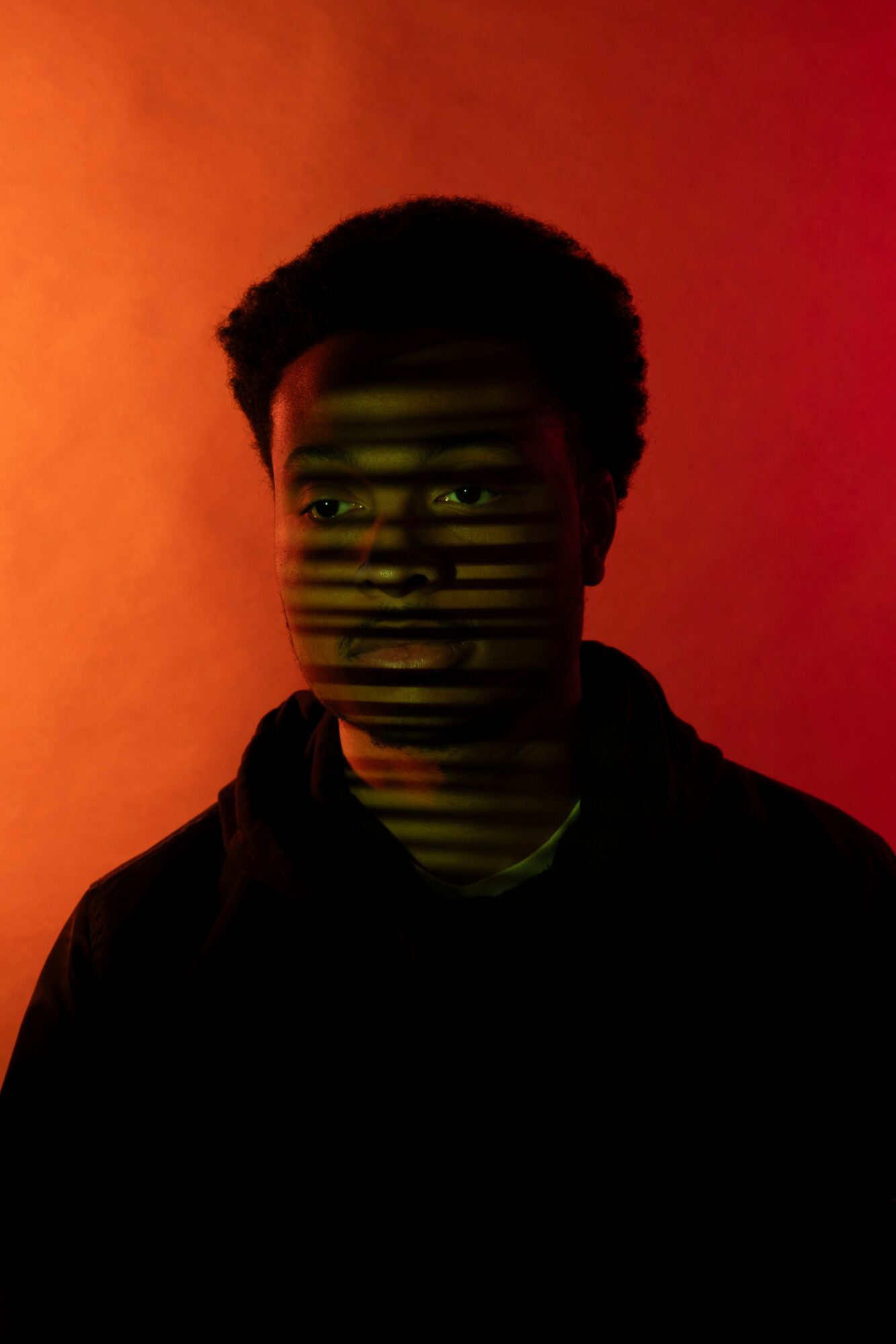 Person with short curly hair faces camera against a red-orange background, with horizontal green lines across face.