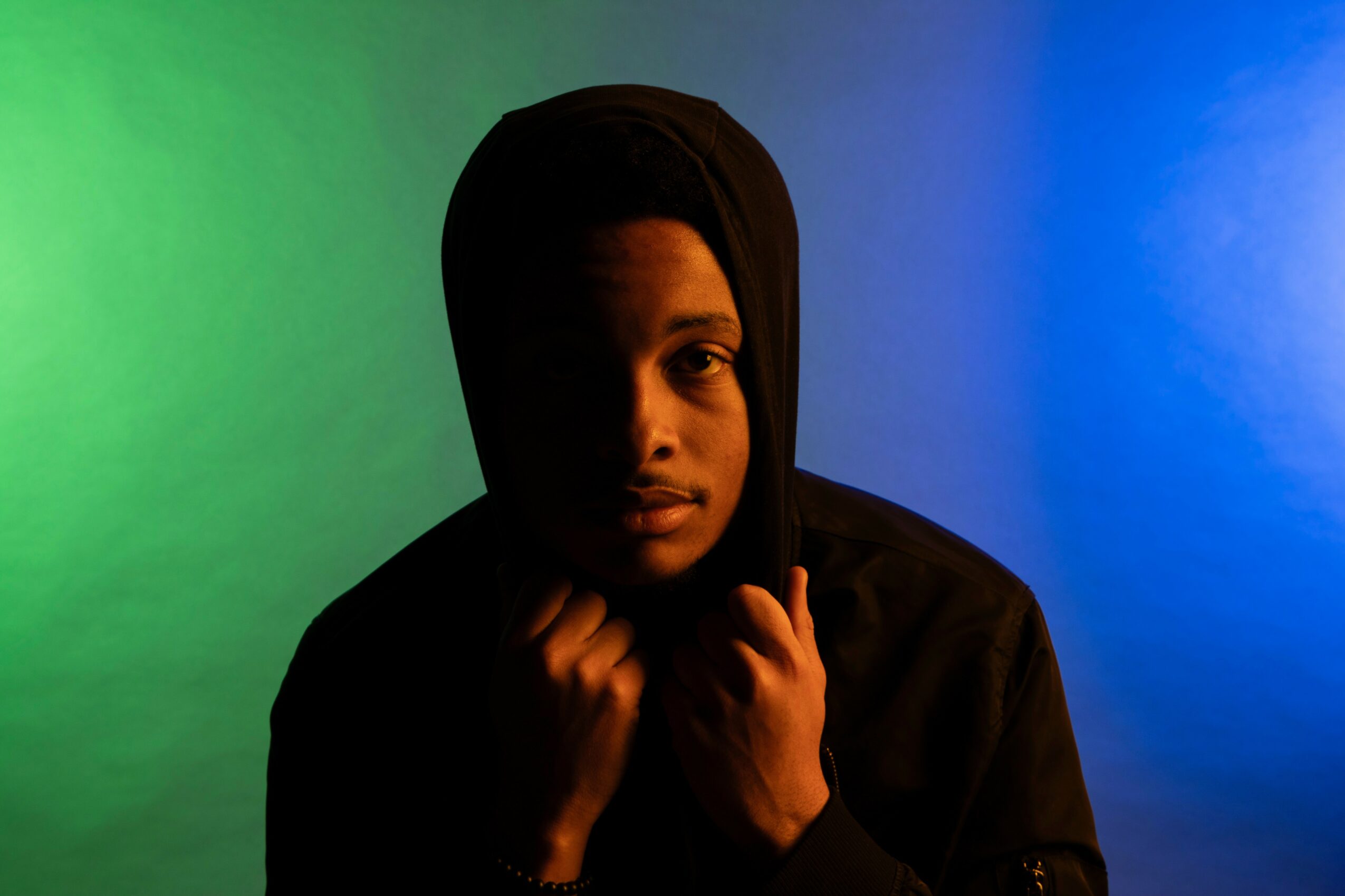 Person wearing a hooded garment, holding their hands near their face, with a green and blue gradient background.