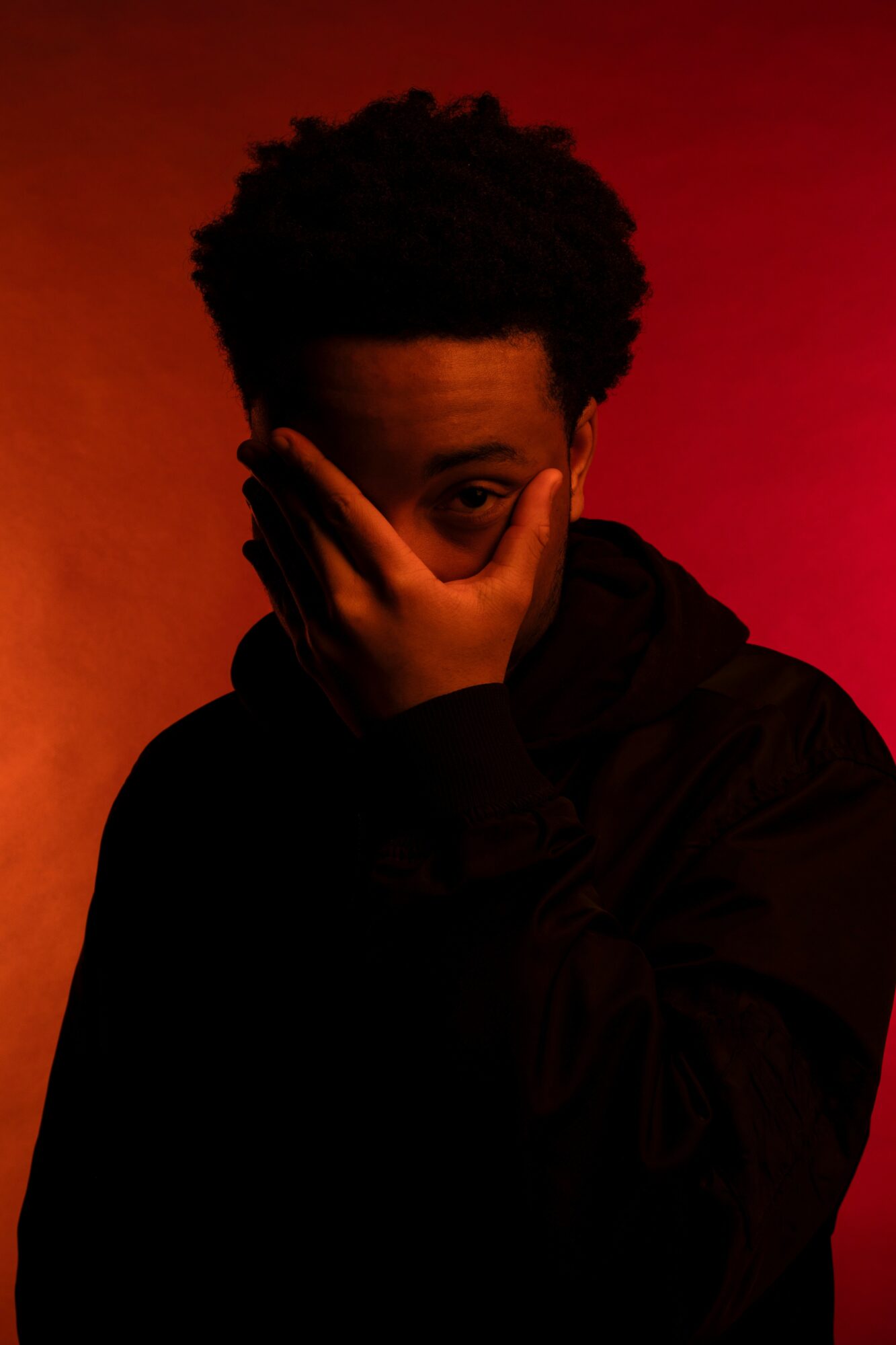 Person with hand covering part of face, dark clothing, background with red and orange gradient