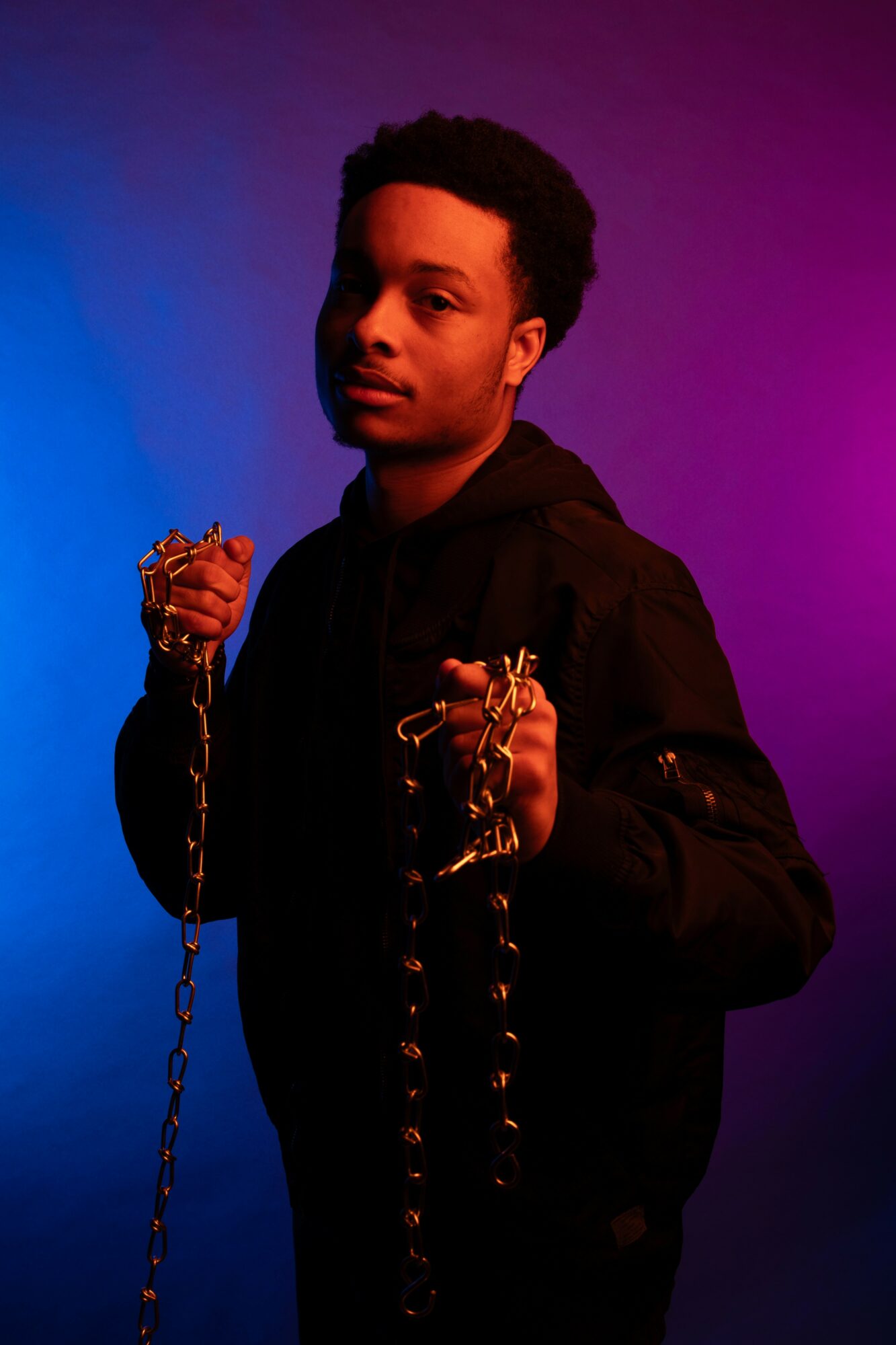 Young person holding chains, standing against a colorful gradient background, looking at the camera with a serious expression.