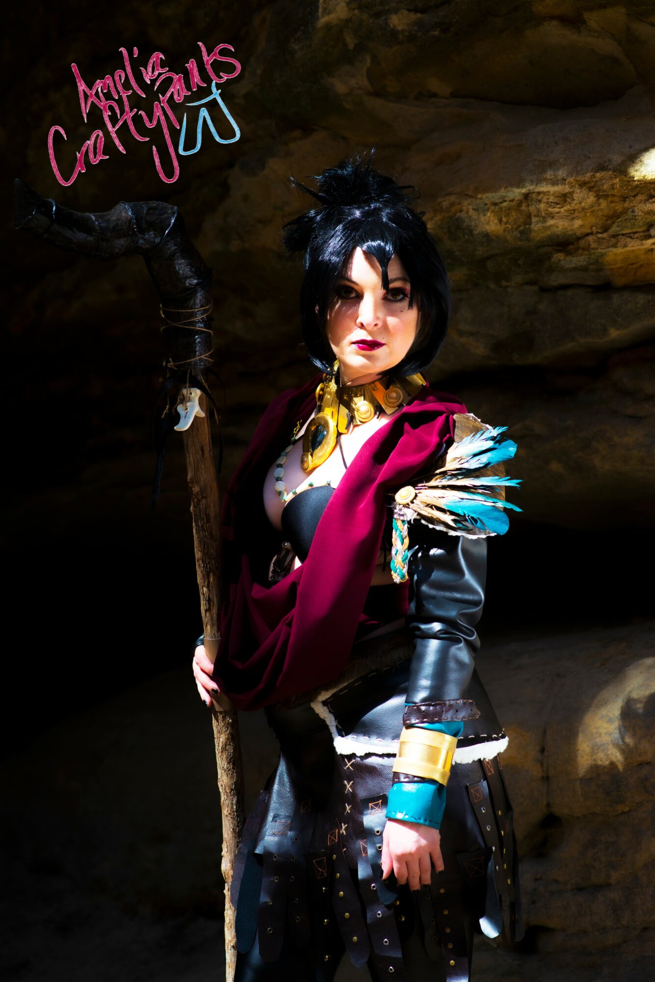 Person in costume with black hair, colorful accessories, and a staff, standing in a rocky environment.