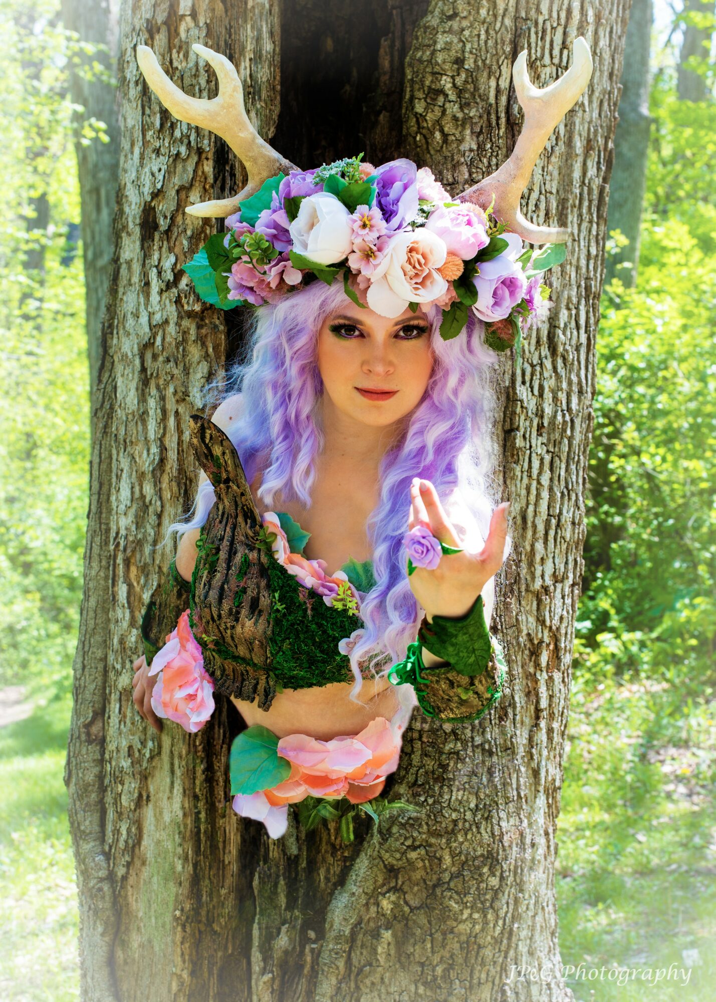 Person with purple hair wearing a floral and antler headpiece, peeking through a tree trunk in a forest.