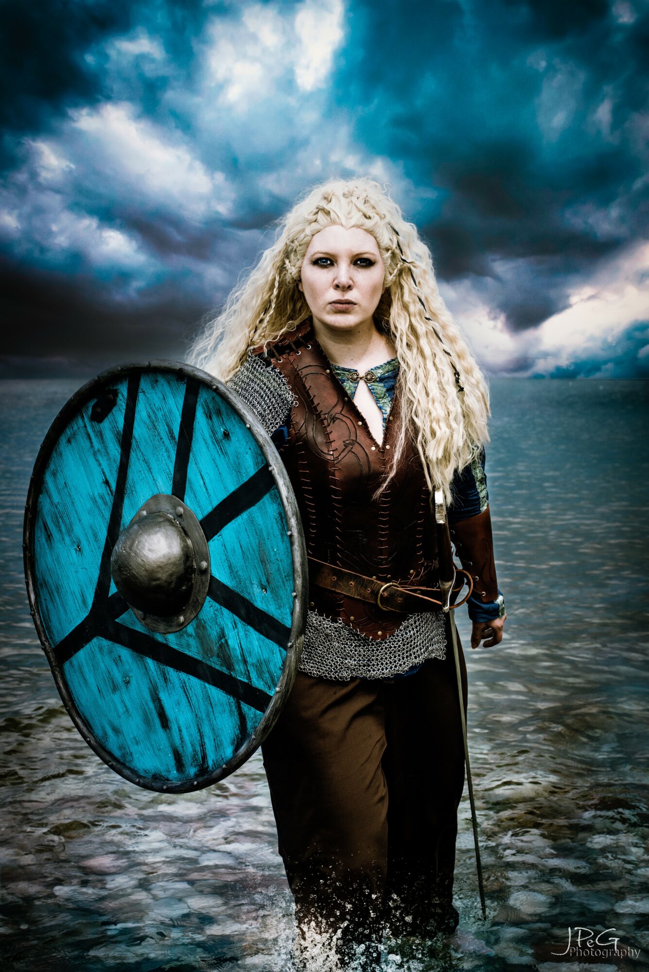 Woman with long curly blonde hair holding a shield, standing in water with stormy sky background.
