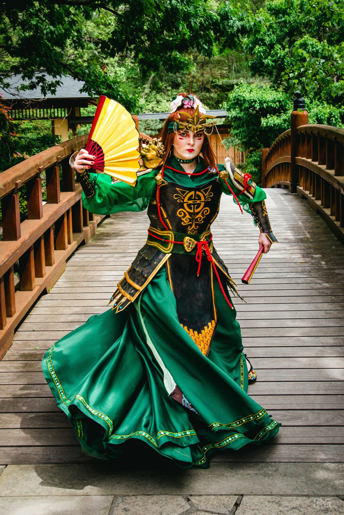 Person in elaborate green and black costume holding a fan on a wooden bridge surrounded by trees.