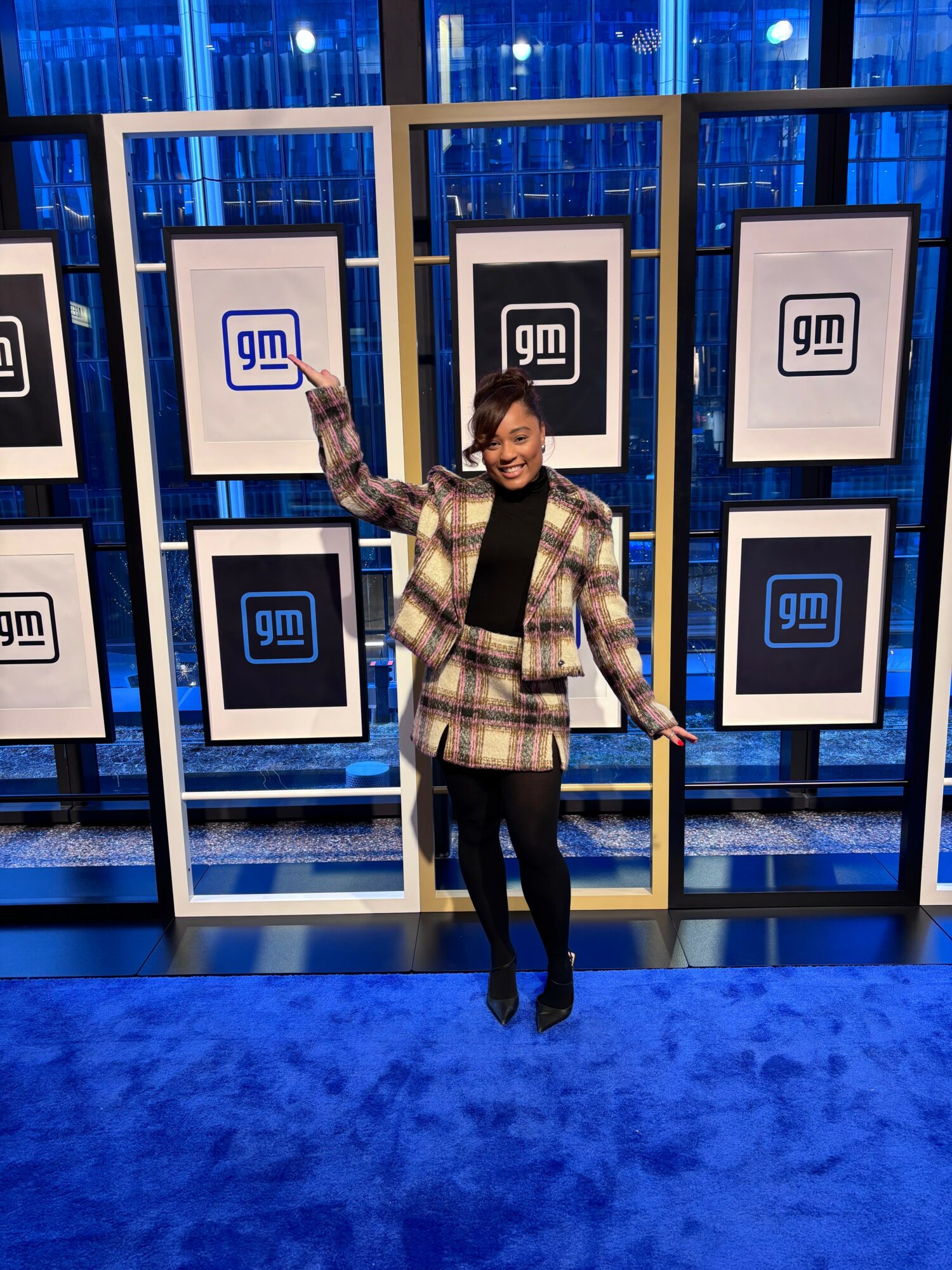 Person standing in front of a display with multiple GM logos, smiling, wearing a plaid jacket and black pants.