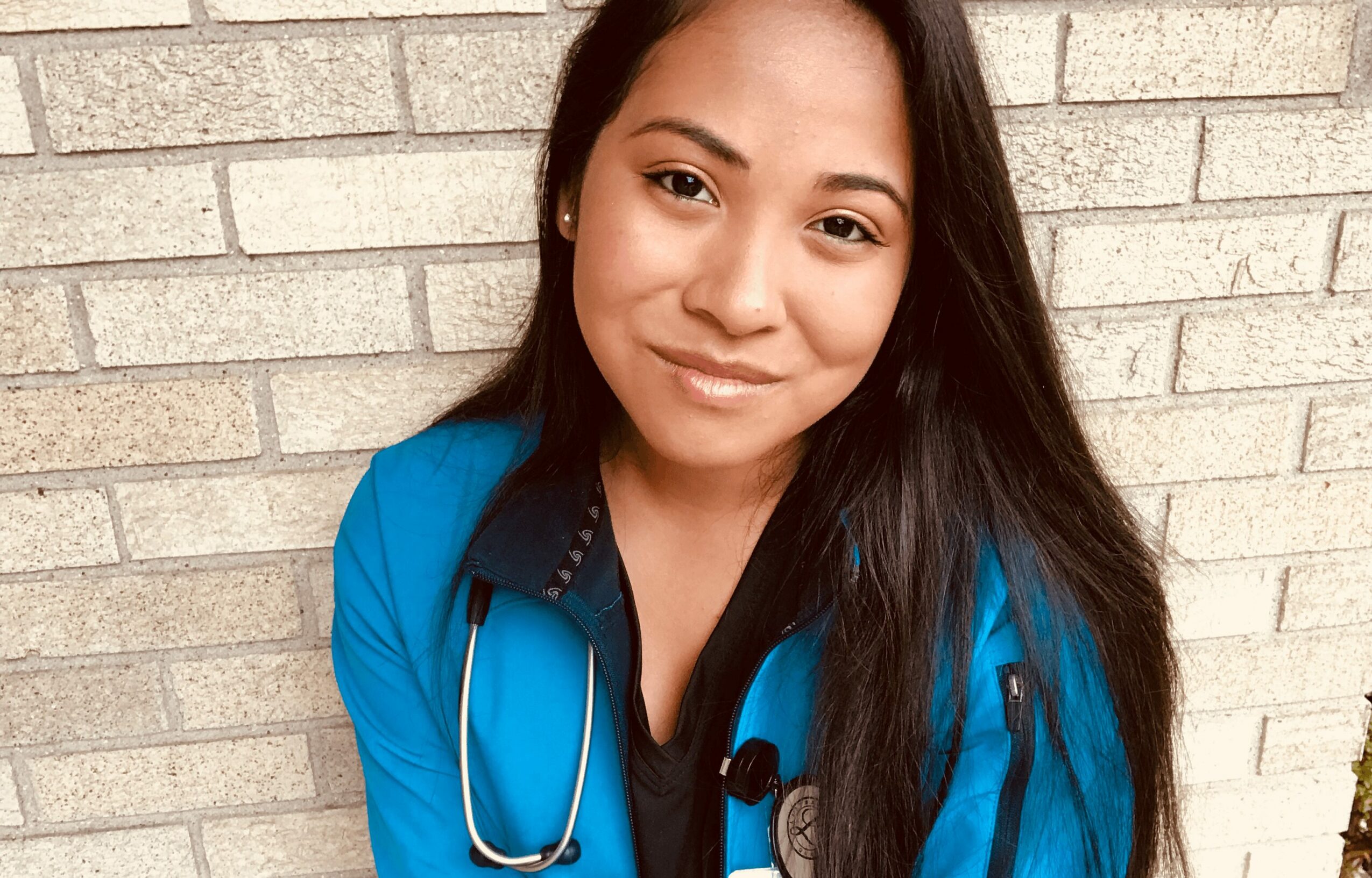 Young woman with long dark hair smiling, wearing a blue jacket and stethoscope, standing against a brick wall.