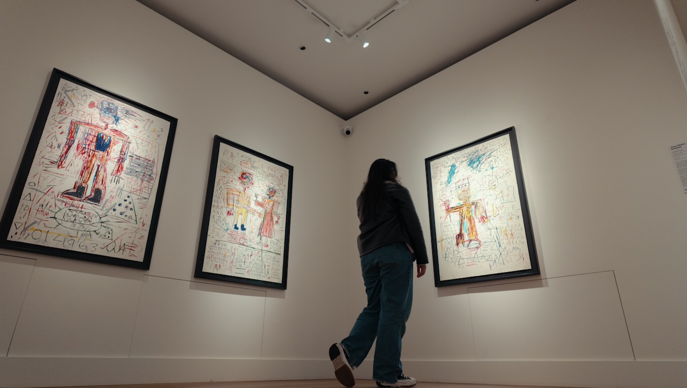 Person viewing three framed artworks on a white gallery wall, ceiling lights overhead, person with long dark hair wearing a dark jacket and jeans, gallery space with high ceiling.