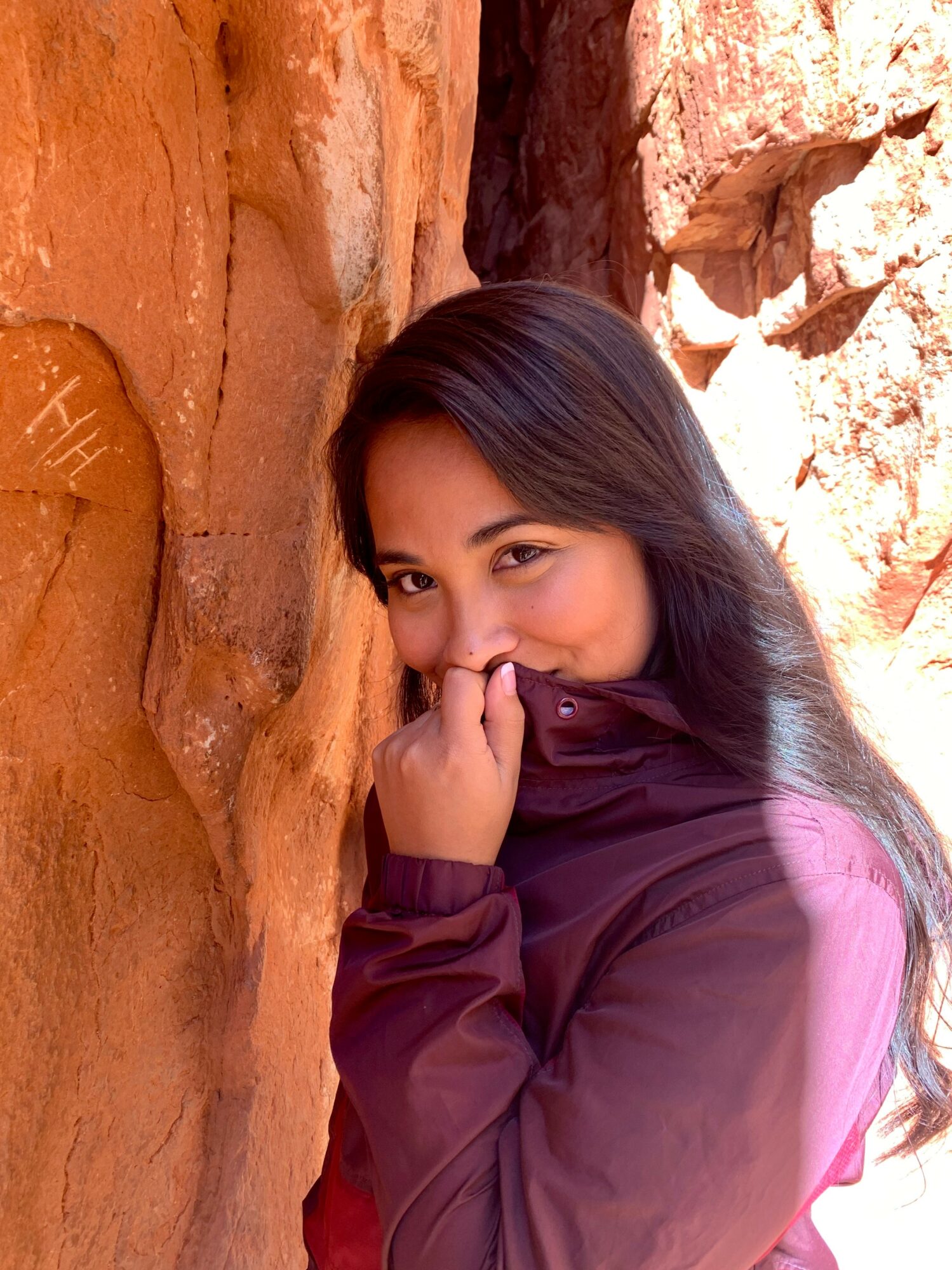 Young woman with long dark hair partially covering her face, wearing a purple jacket, standing against a rocky background.