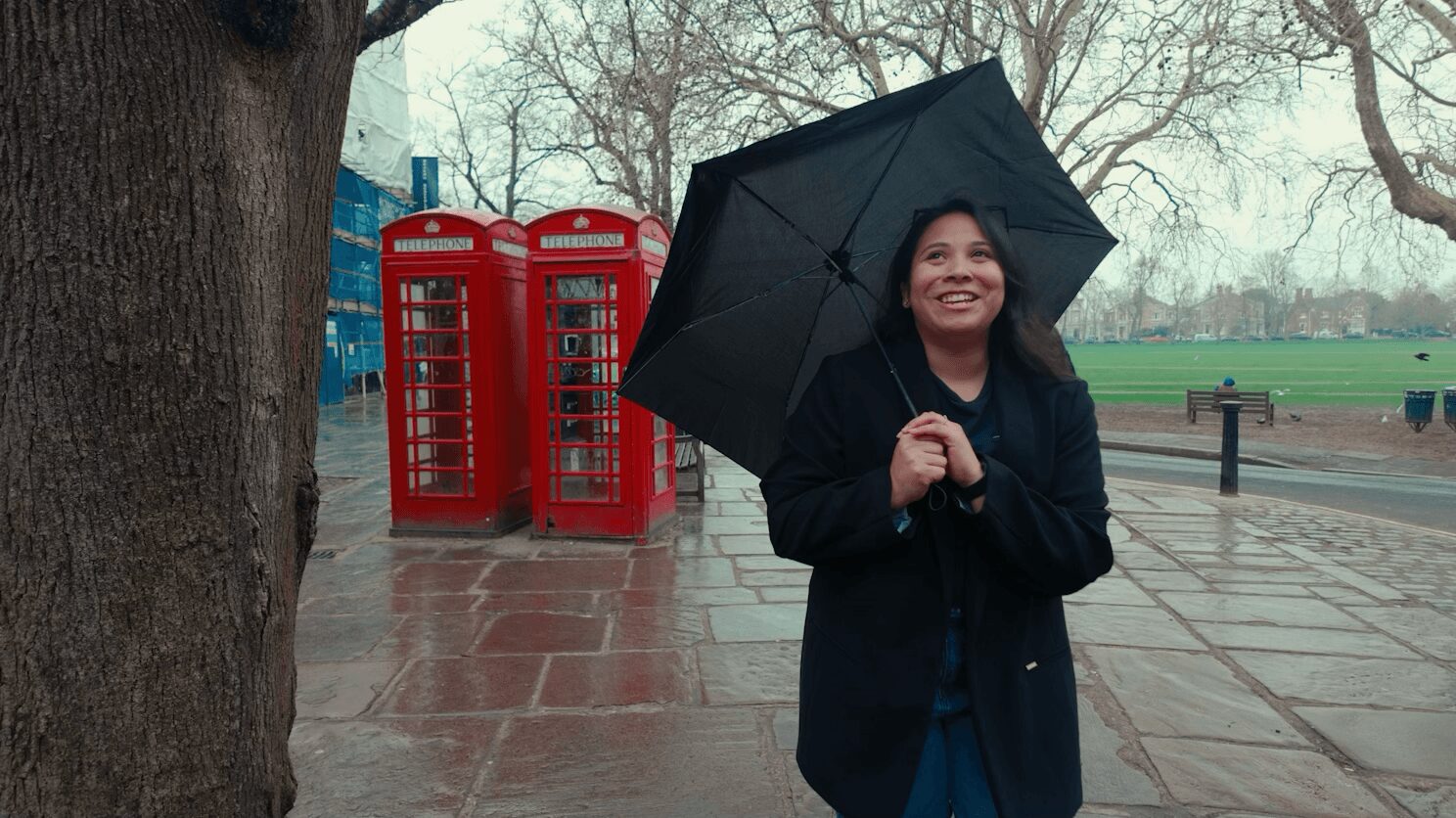 Woman holding an umbrella smiling outdoors on a rainy day, with red phone booths in the background.