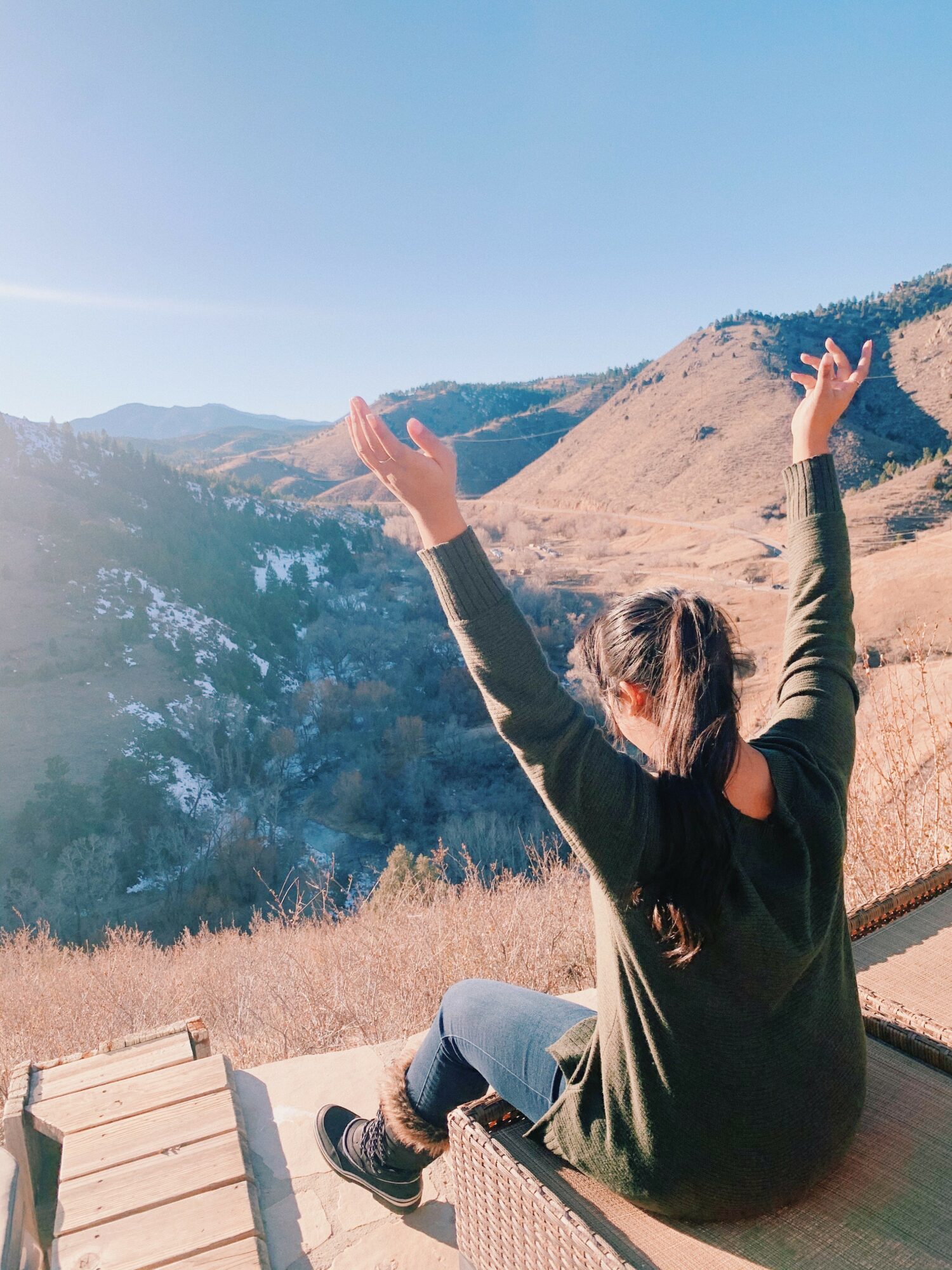 Person sitting outdoors with arms raised, mountains in background, sunny sky, wearing dark clothing, hair tied back, sitting on a bench.