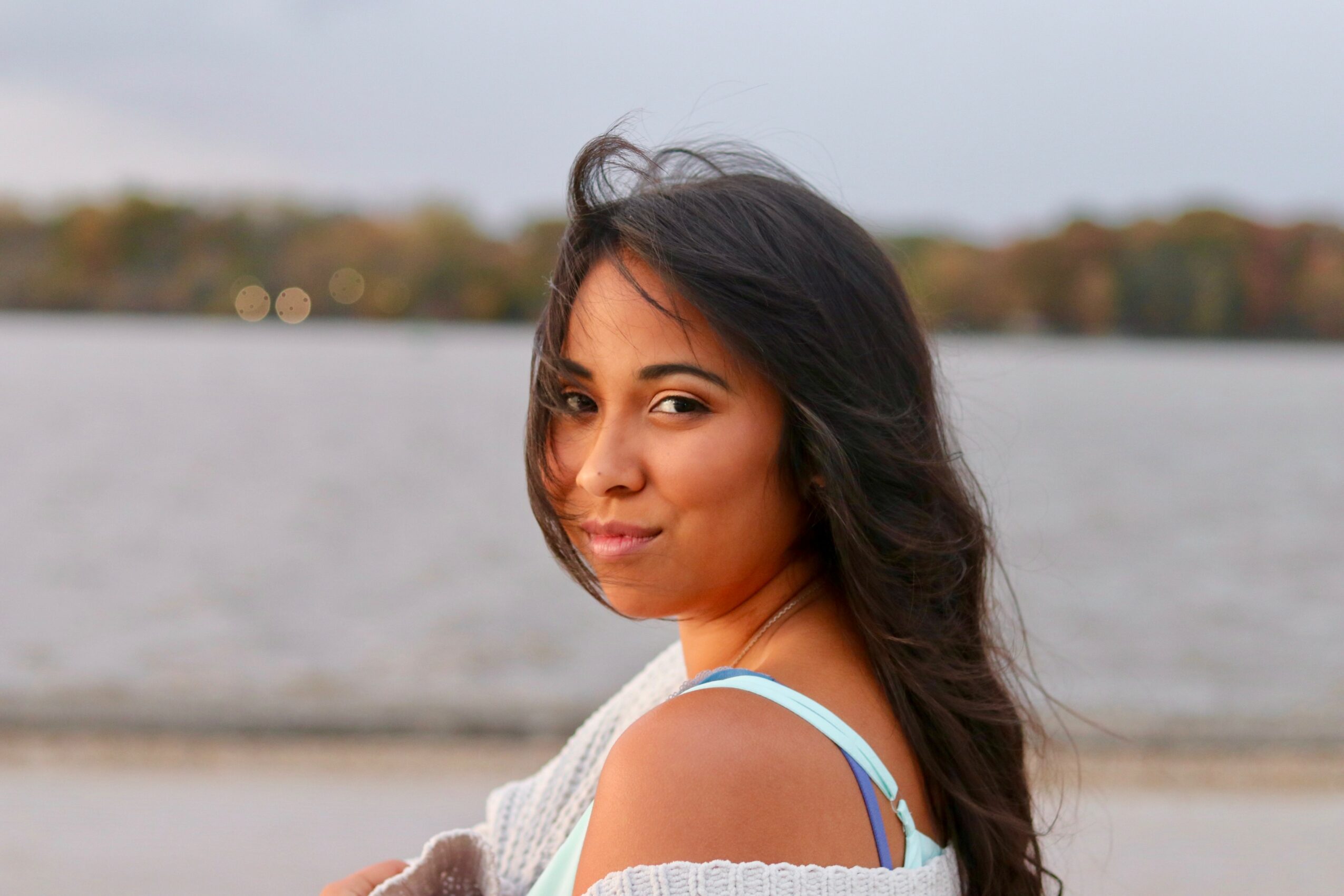 Young woman with long dark hair smiling outdoors near a body of water, wearing a light-colored top and blue straps.