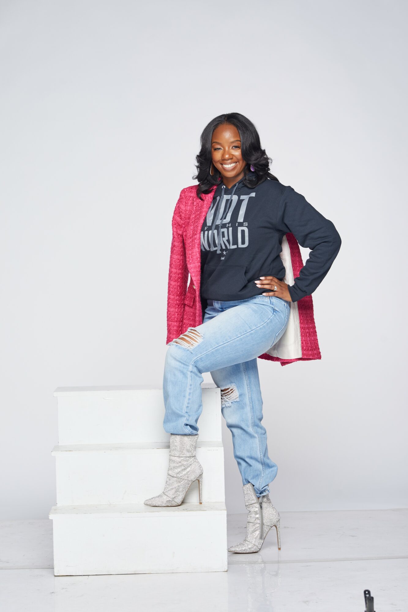 Woman standing on white steps, smiling, wearing a black hoodie, pink jacket, ripped jeans, and high-heeled boots.