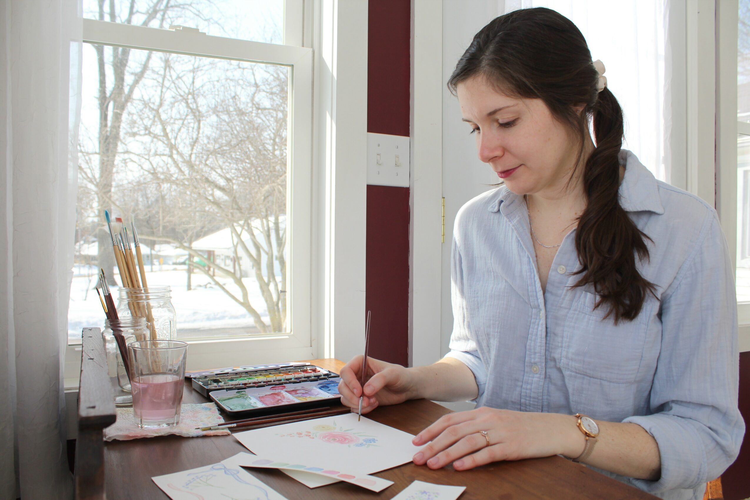 Woman sitting at a desk, looking at papers with a window showing trees outside behind her.