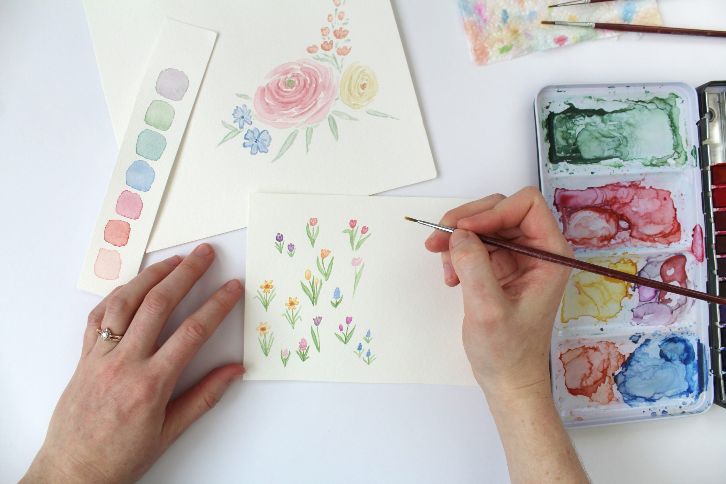 Person painting floral watercolor on paper with watercolor palette nearby, hand holding brush, colorful artwork in progress.