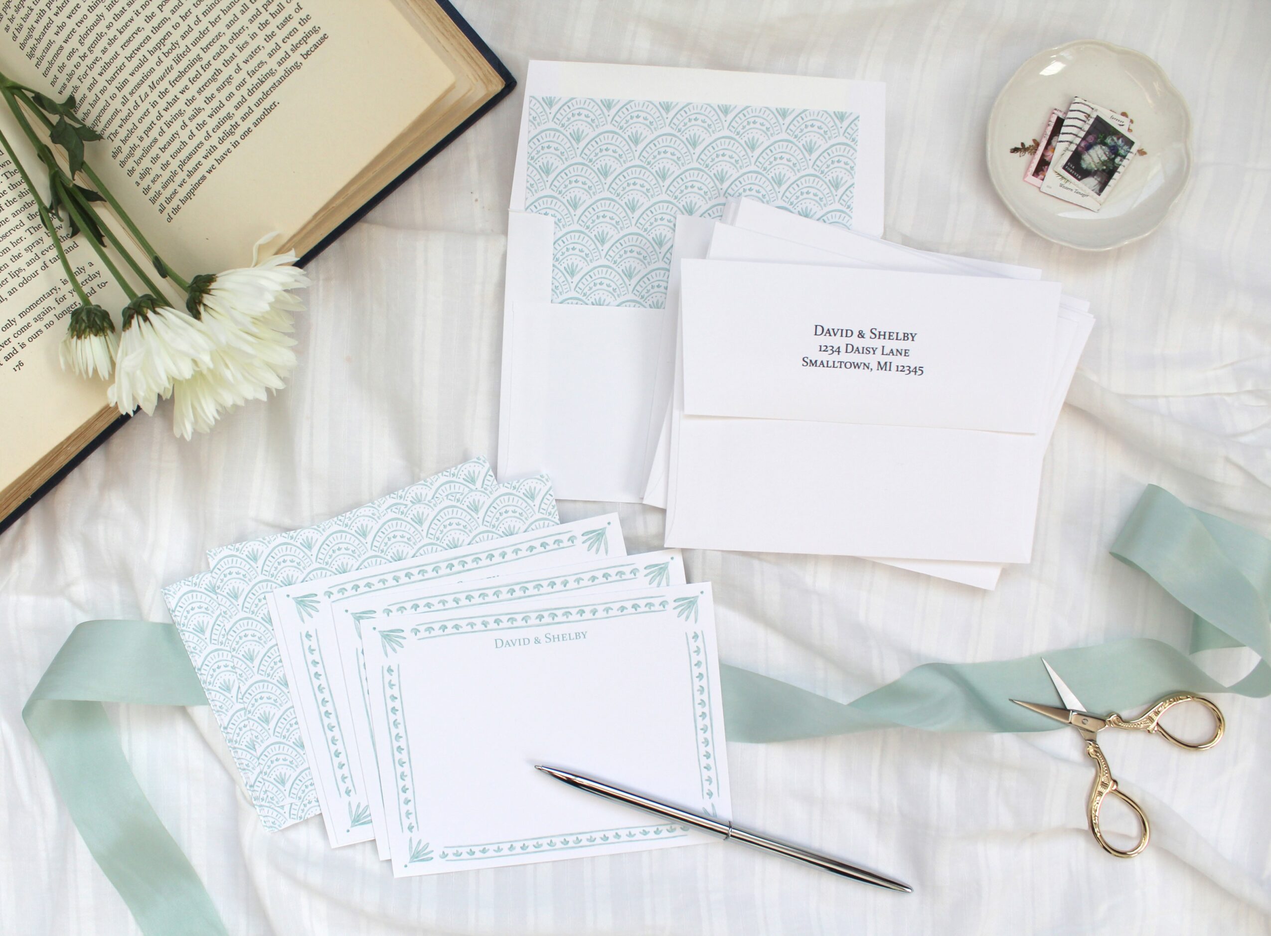 Stationery items including envelopes, cards, a pen, scissors, and a book on a white fabric surface with flowers and decorative objects.