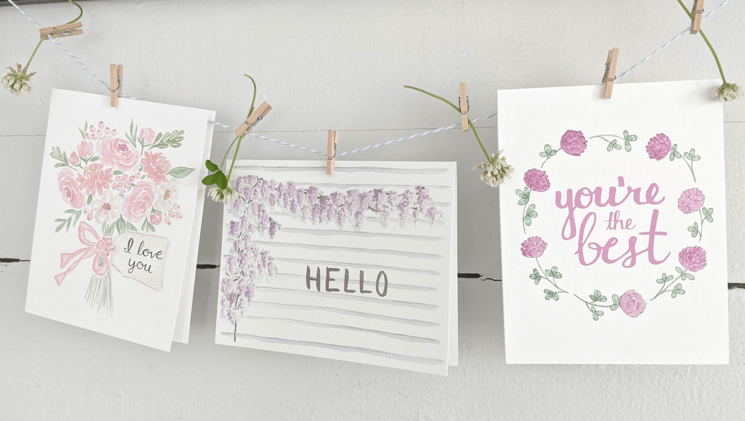 Three greeting cards hanging on a string with clothespins, decorated with flowers and messages, against a white background.
