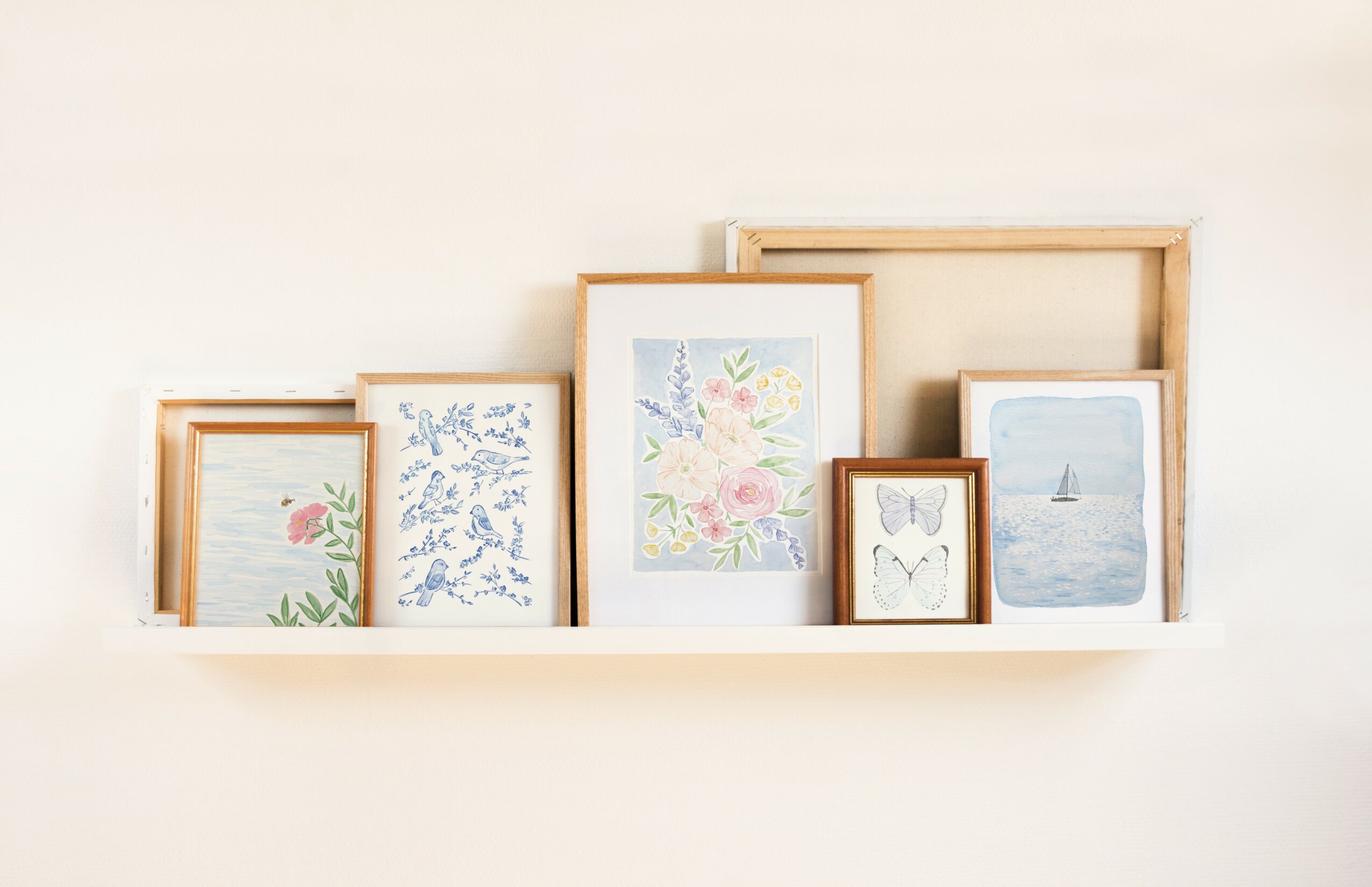 Six framed pictures on a white shelf, including floral, abstract, and landscape images, against a plain wall.