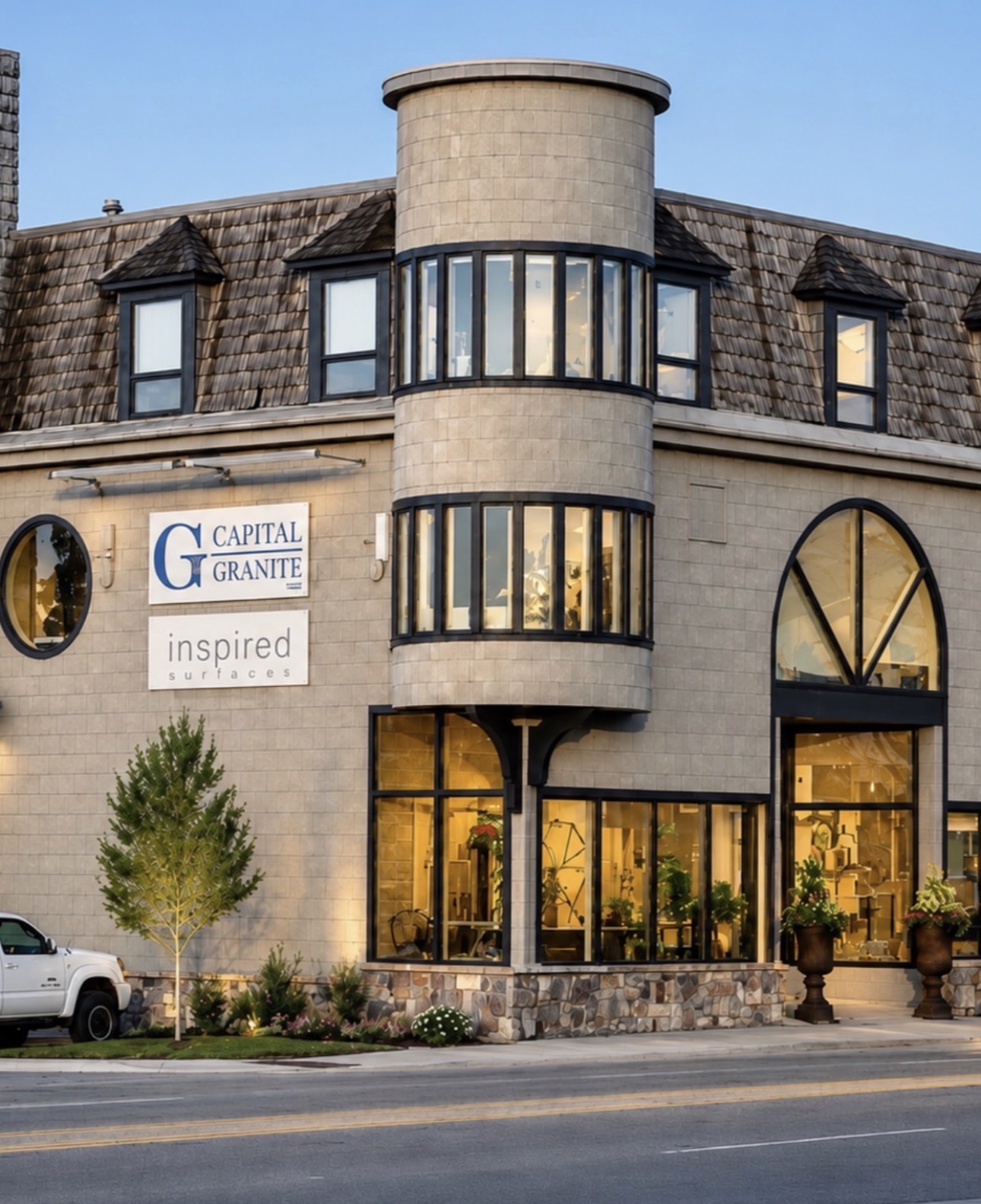 Building with rounded tower, large windows, and a sign reading 'CAPITAL GRANITE inspired', with a tree and parked car in front.