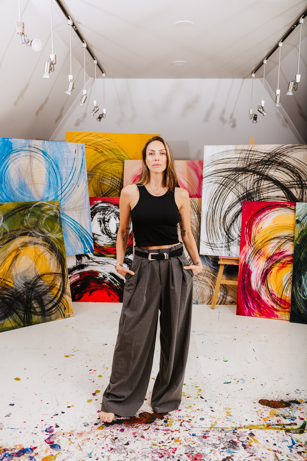 Woman standing in front of colorful abstract paintings in an art gallery, wearing a black top and wide gray pants.