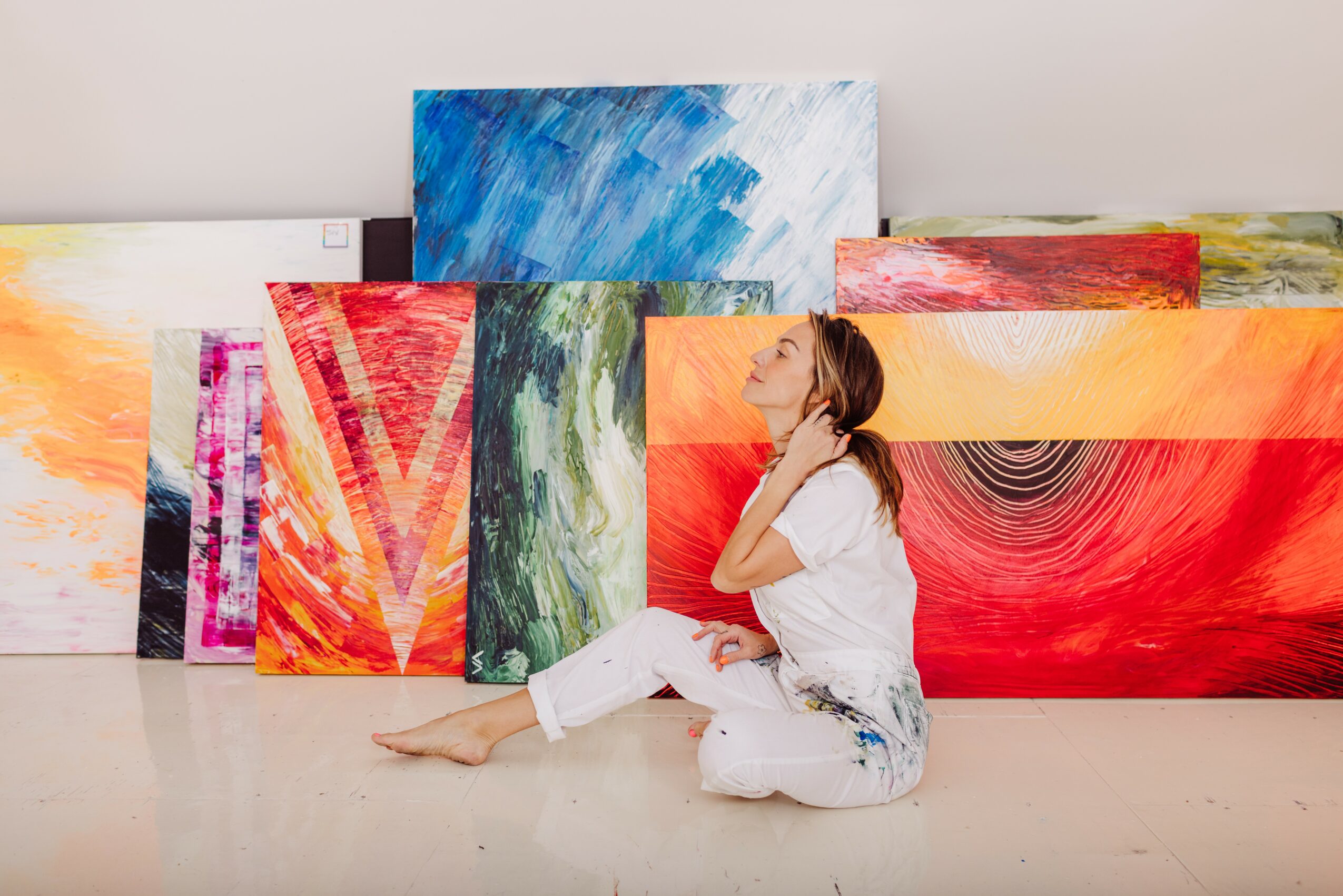 Woman sitting on floor surrounded by colorful abstract paintings, leaning back with hand on neck, in an art gallery.