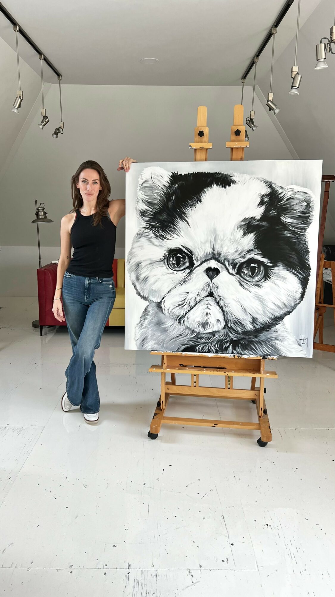 Woman standing next to a large black and white dog portrait on an easel in a studio.