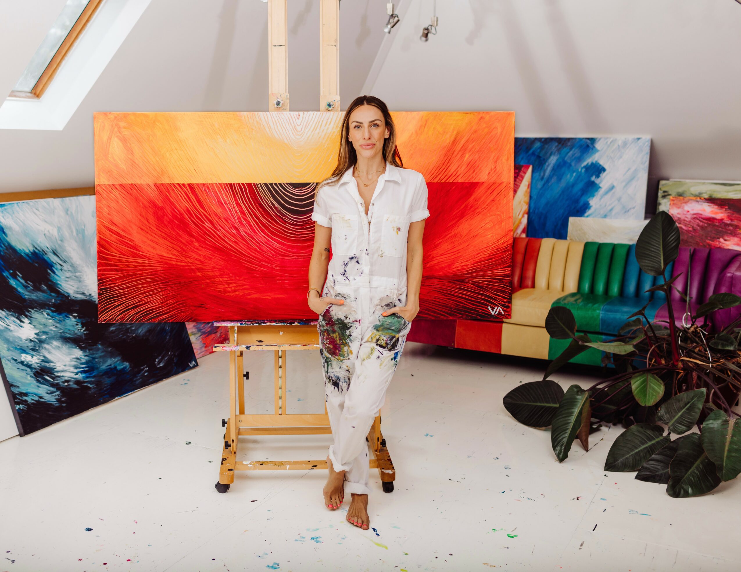 Woman standing in art studio with colorful paintings, plants, and furniture, wearing white clothes with paint splatters.