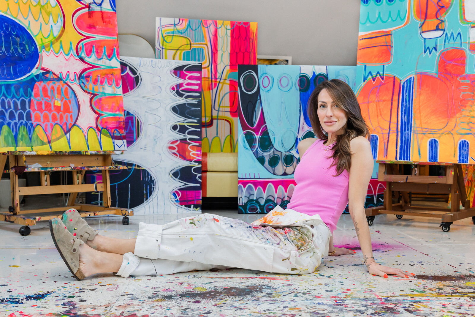 Young woman sitting on the floor in front of colorful abstract paintings, smiling, with paint splatters on the floor.