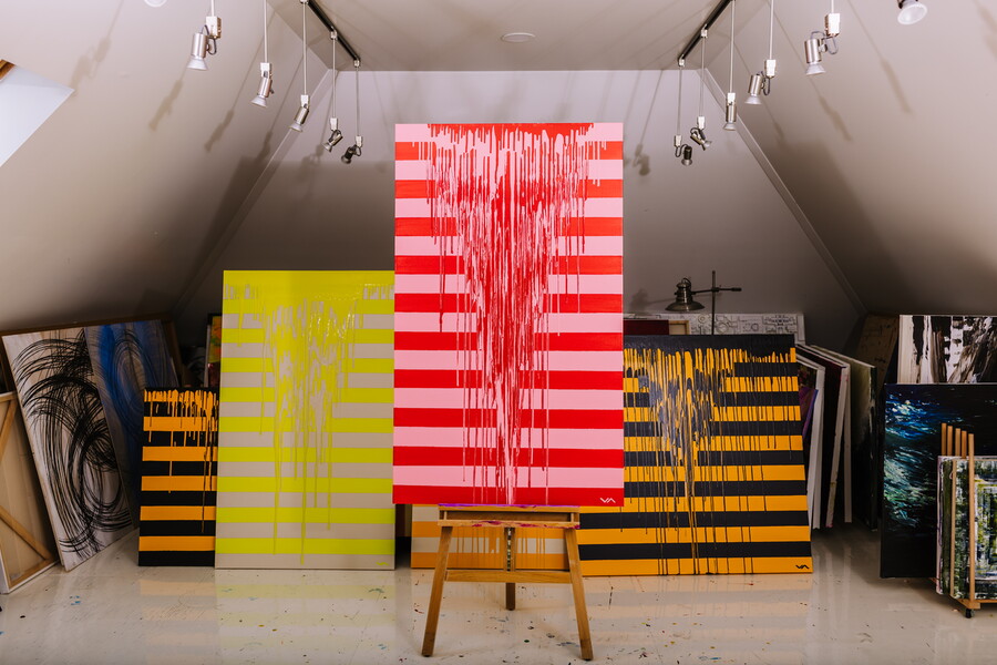 Gallery with colorful striped artwork on display, yellow and black striped pieces, and a wooden stool in front, under angled ceiling lights.