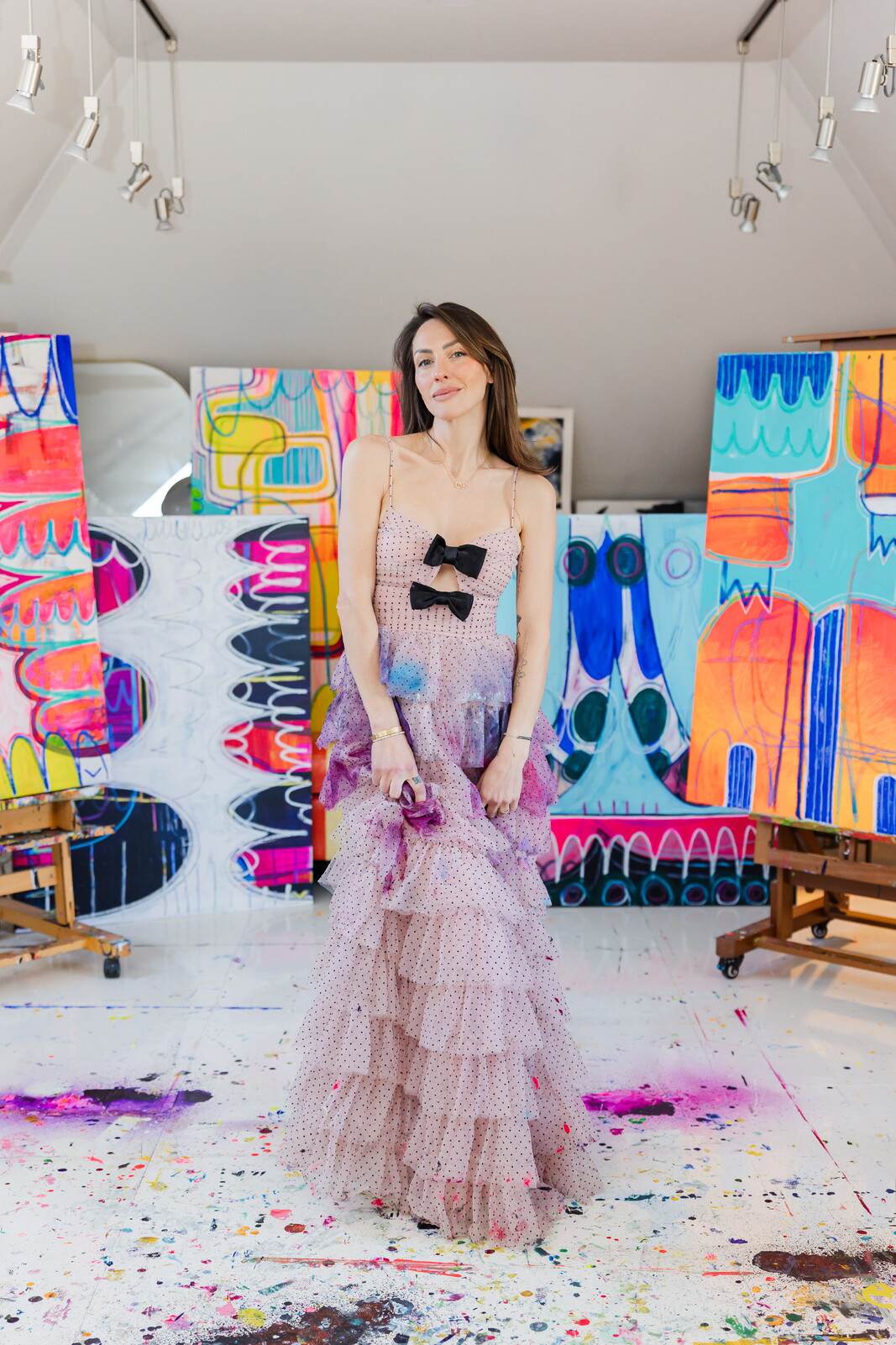Young woman in a layered pink dress with black bow, standing in front of colorful abstract paintings, in an art gallery.