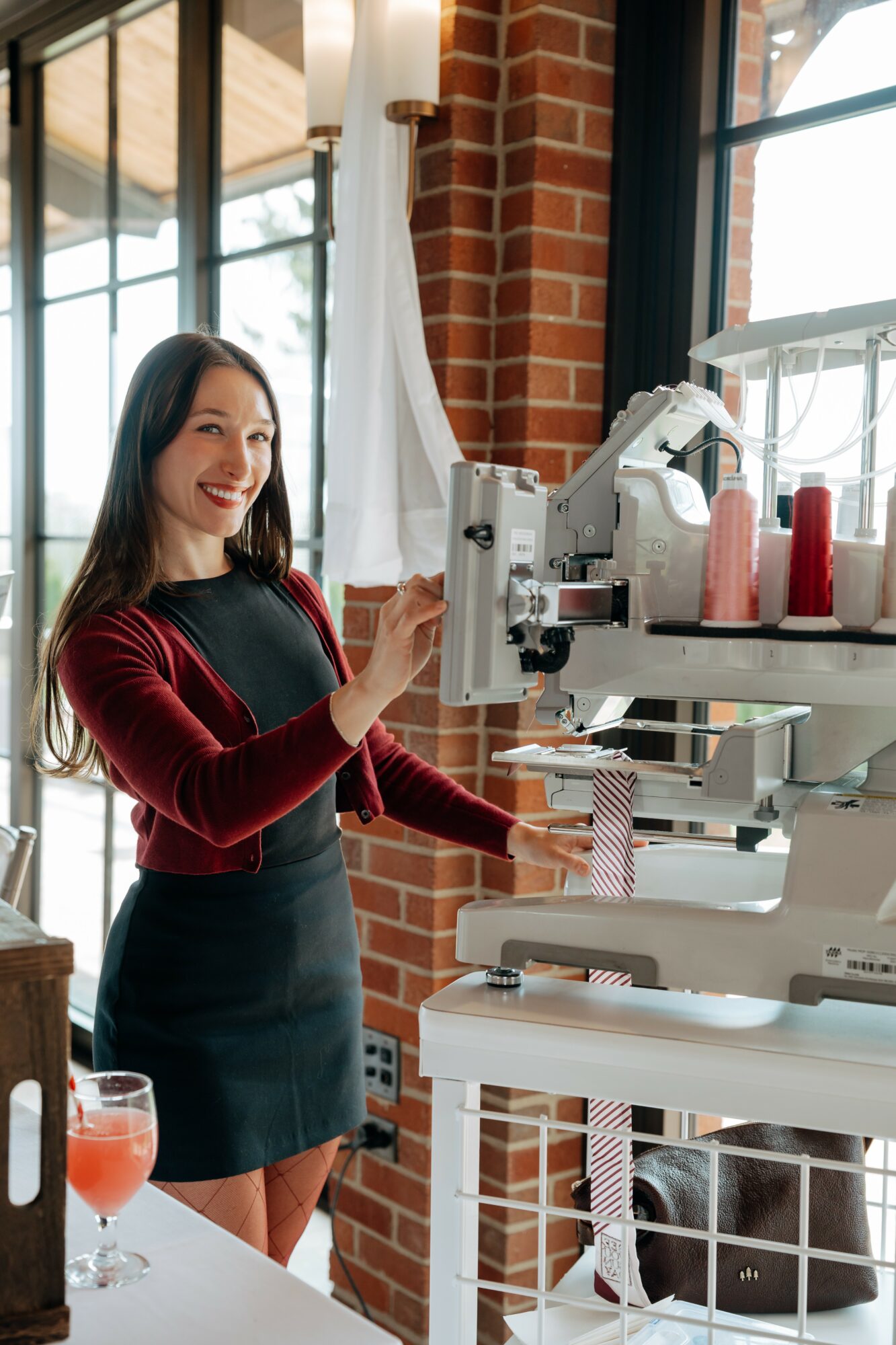 Woman smiling at a machine in a brick-walled, sunlit room with large windows, holding a small object, wearing a red top and black skirt.