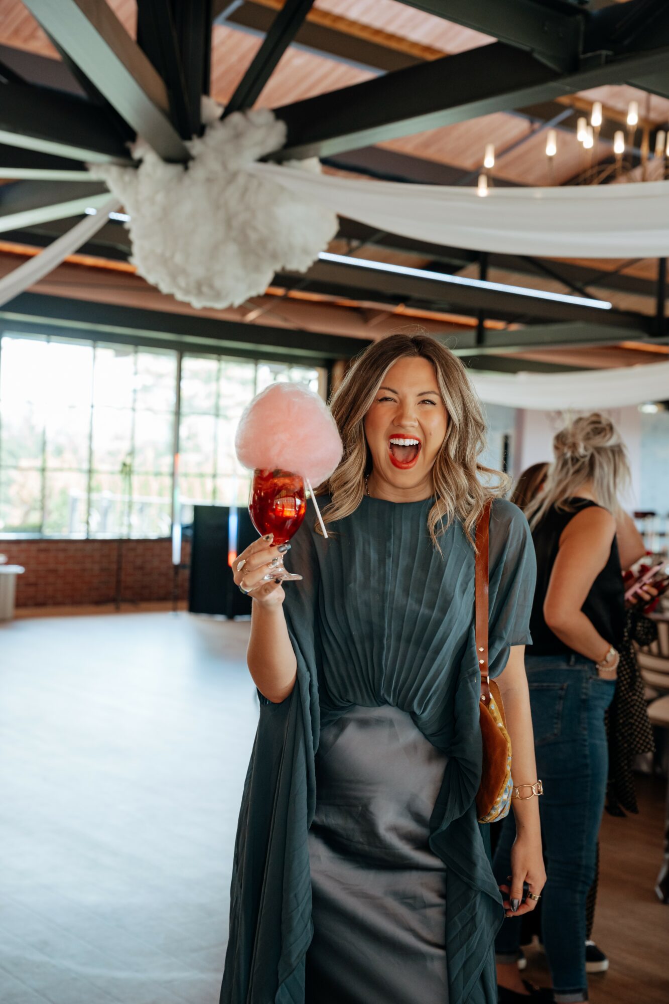 Woman smiling and holding a pink cotton candy at an indoor event, with other people in the background.
