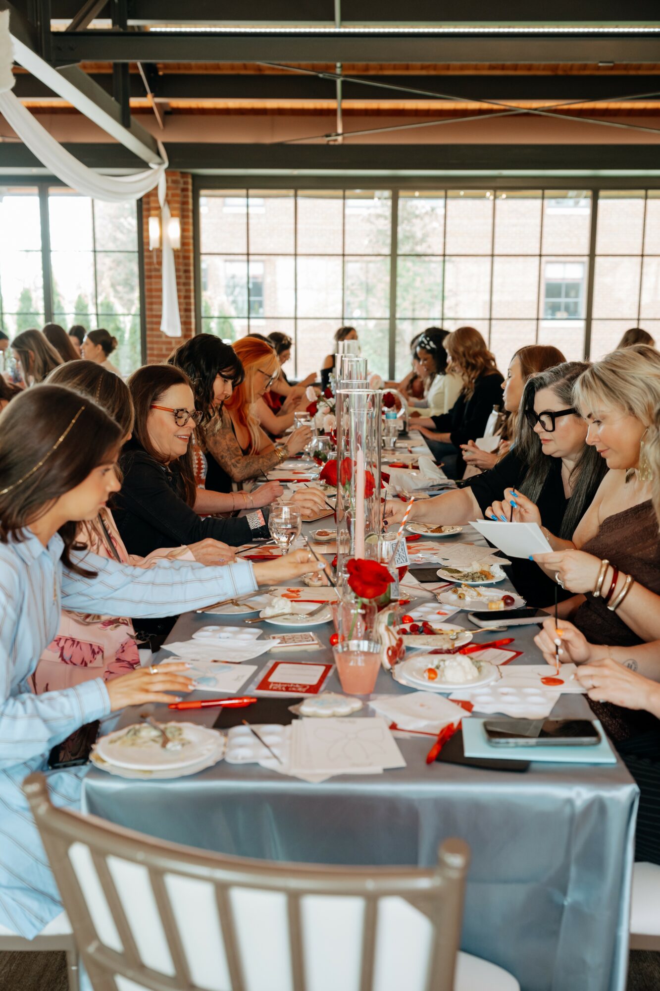 People seated at a long table with flowers and papers, in a bright room with large windows.