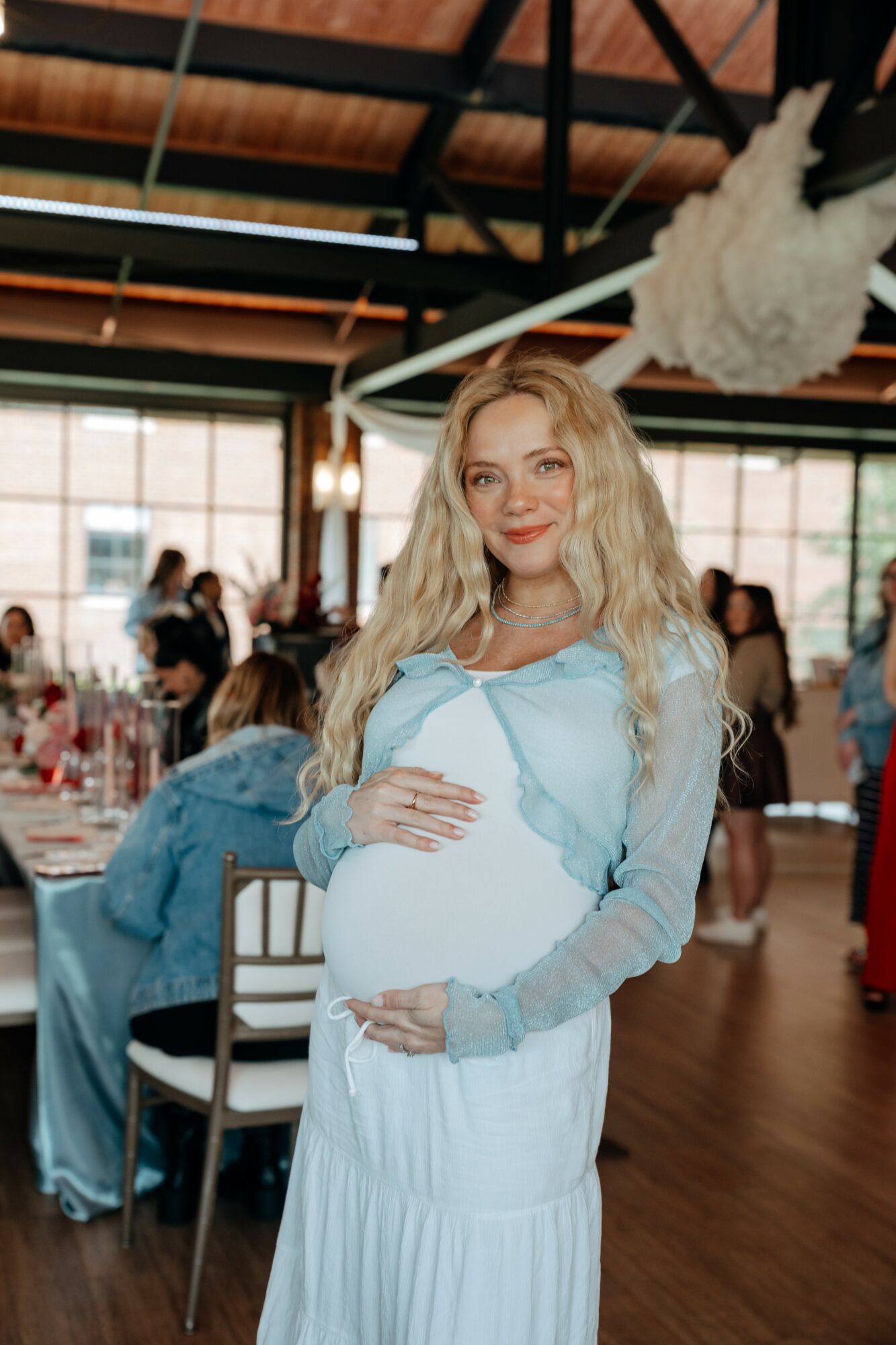 Pregnant woman with long blonde hair smiling, wearing a light blue top and white skirt, in a decorated indoor event space.