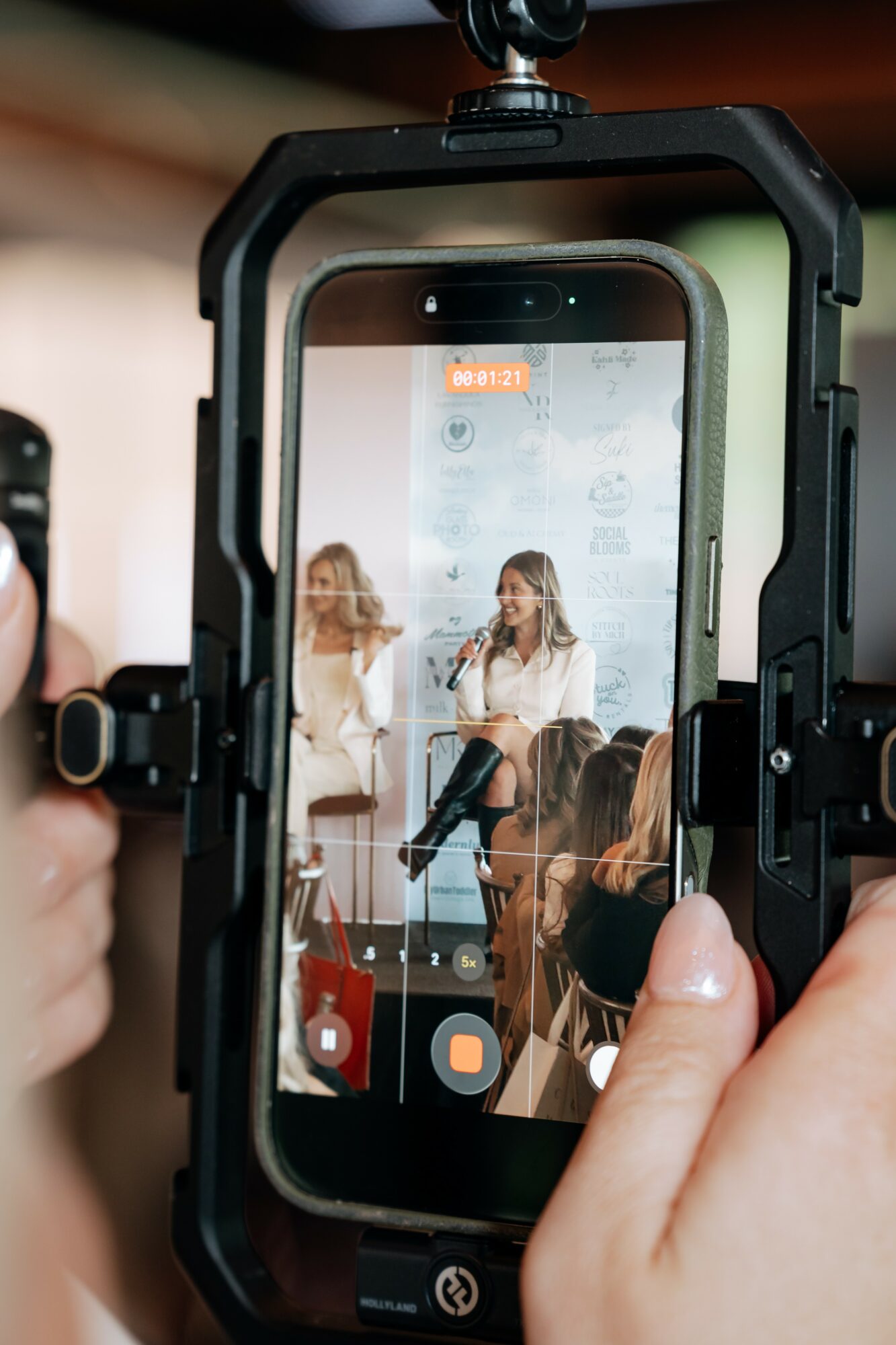 Person recording a woman speaking at a panel discussion with an audience, using a smartphone in a protective case.