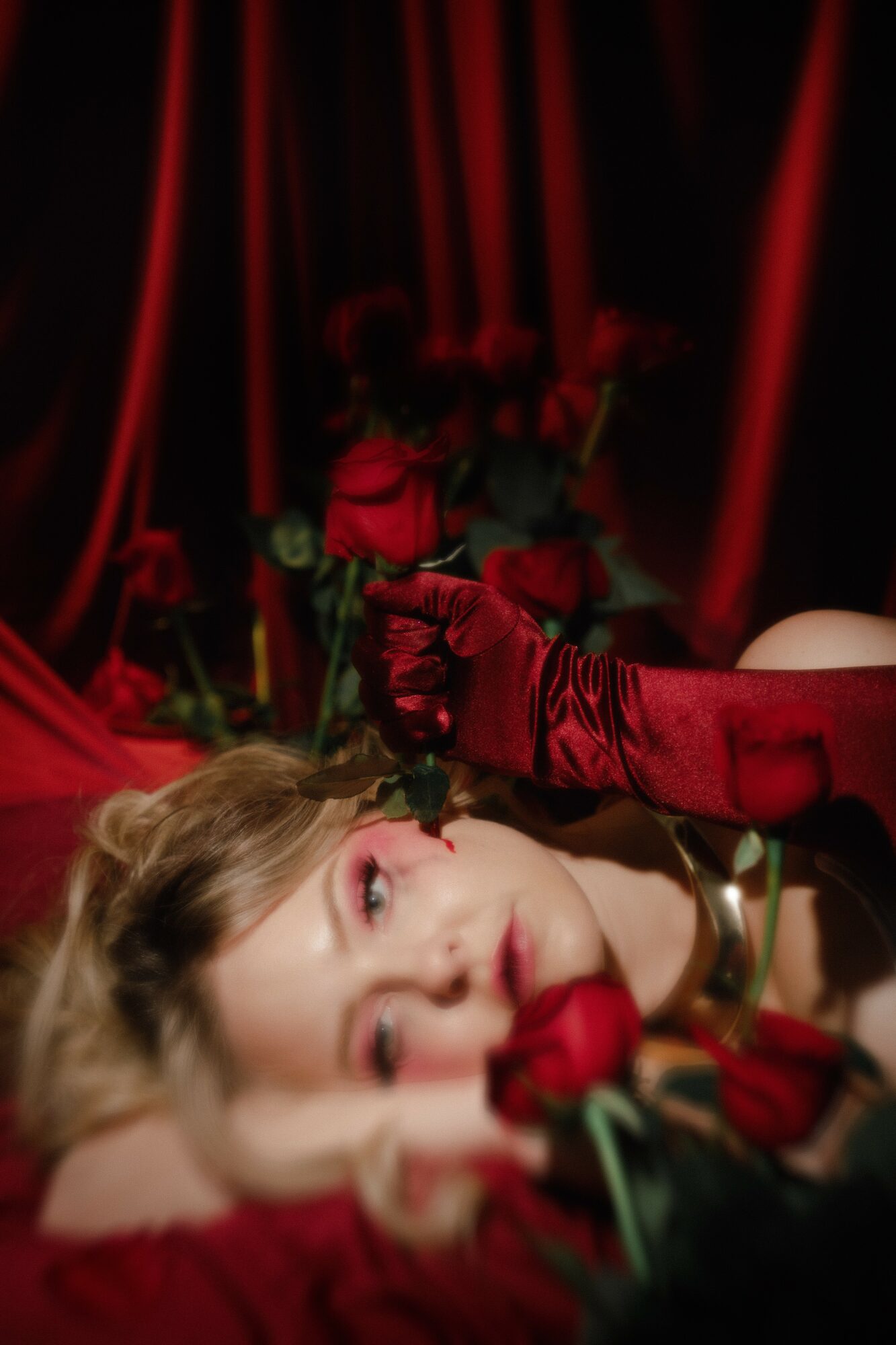 Person lying down with a rose, surrounded by red curtains and roses, wearing red gloves, in a theatrical setting.