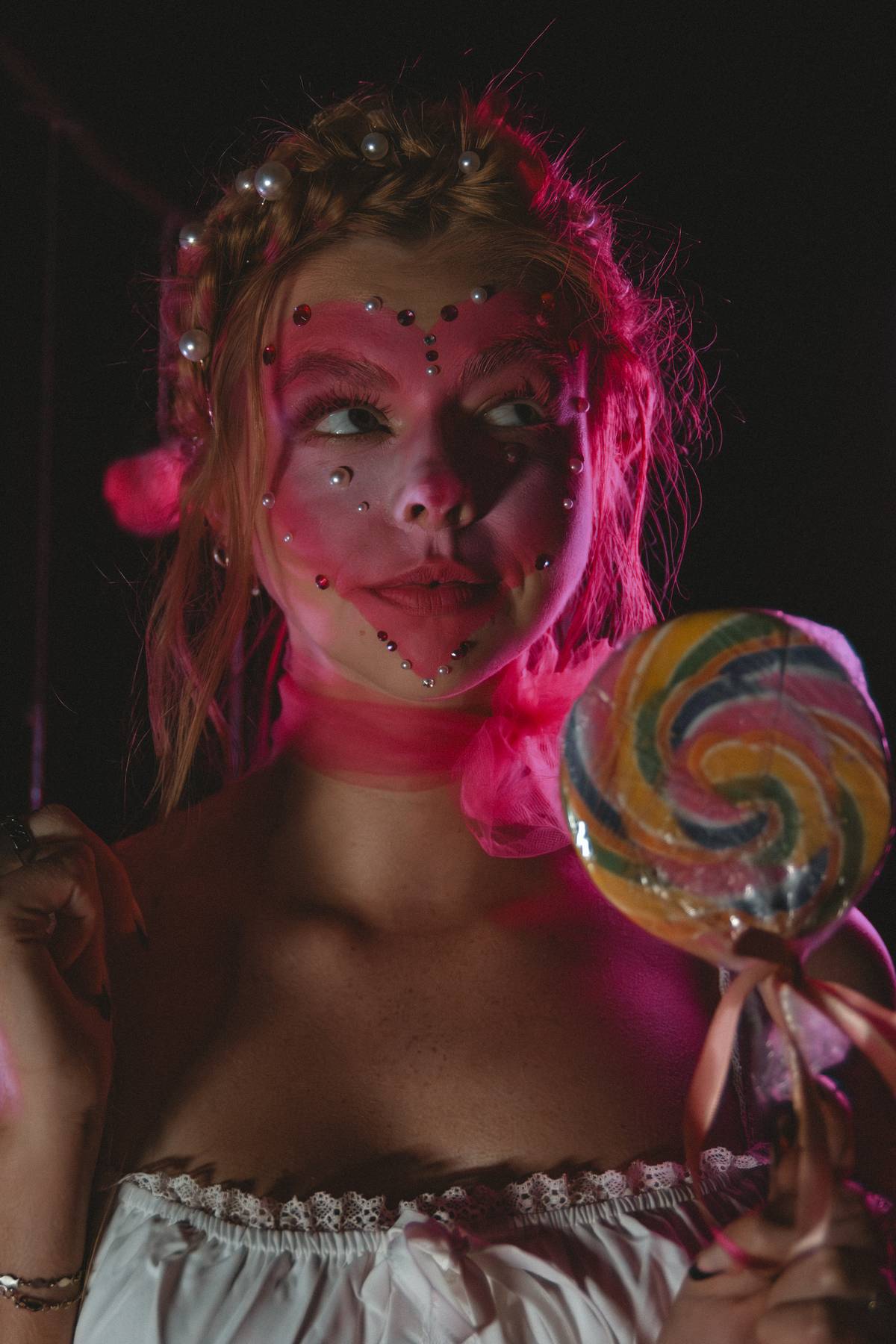 Person with face painted as a clown, holding a colorful lollipop, wearing a ruffled top, with pink hair and makeup.