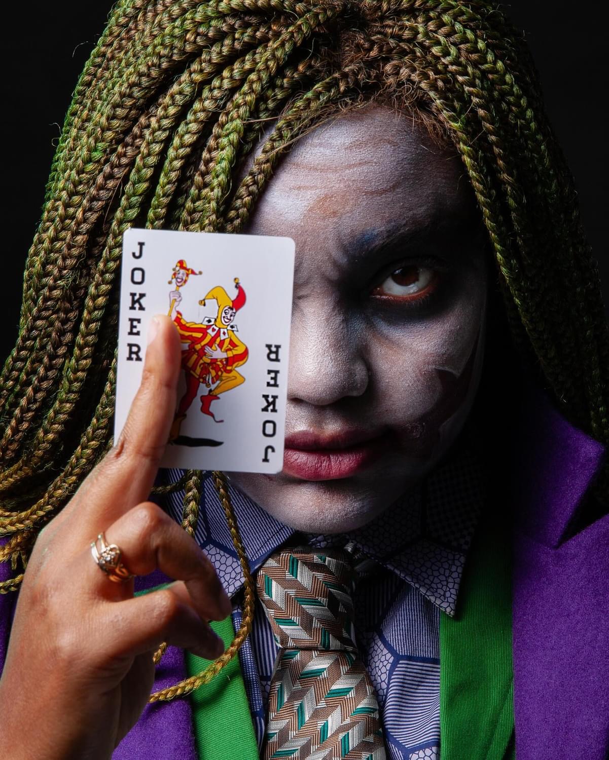 Person with face painted white, holding a Joker playing card in front of their face, with green braided hair and colorful clothing.