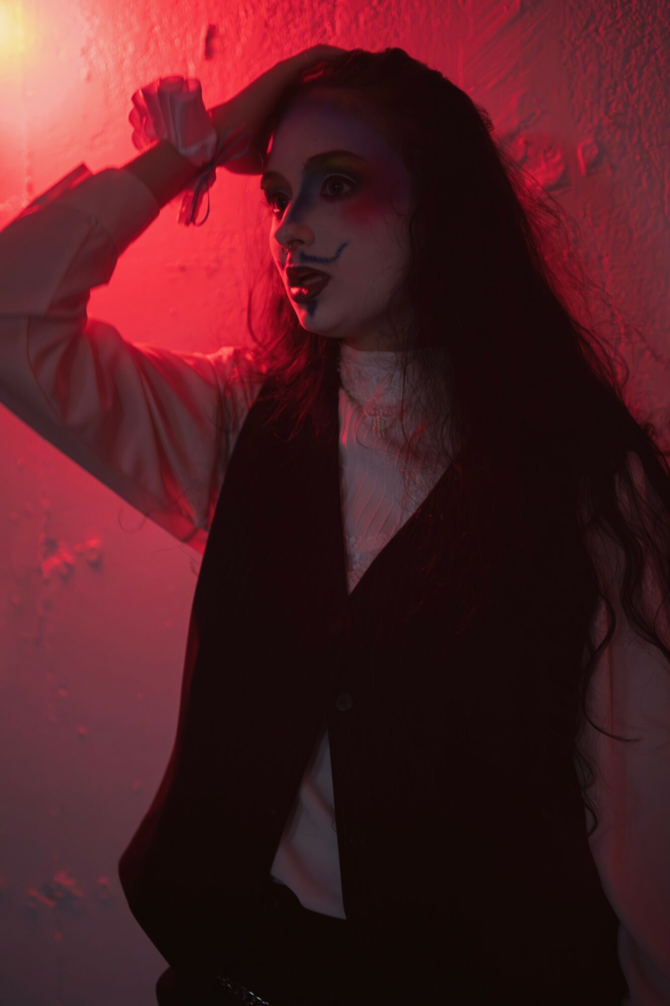 Woman with dark hair and makeup, leaning against a wall illuminated by red light, with her hand on her head.