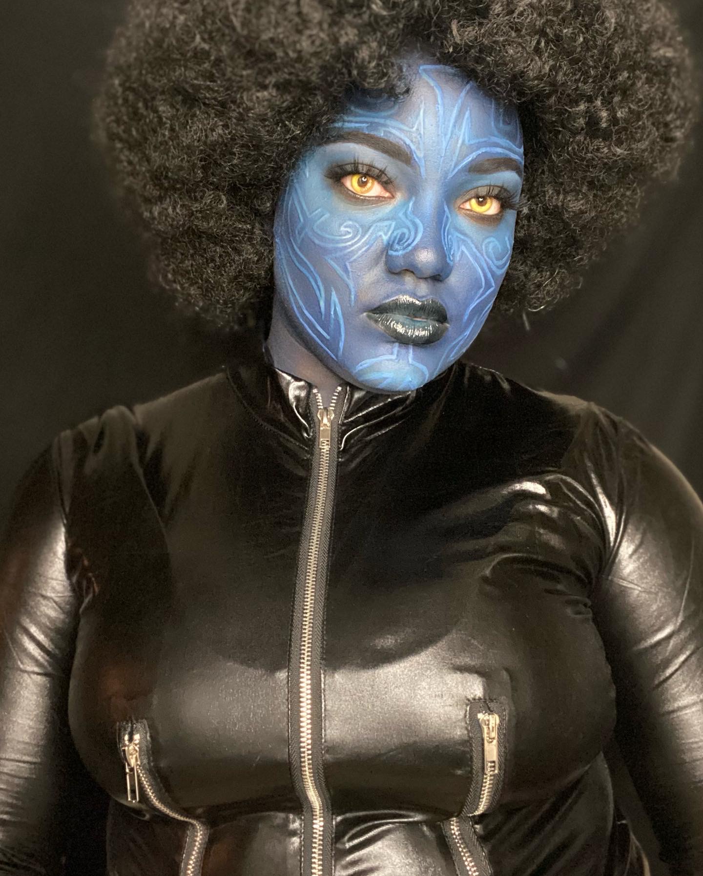 Person with blue face paint, yellow eyes, black lips, wearing a black shiny jacket, and large curly hair.