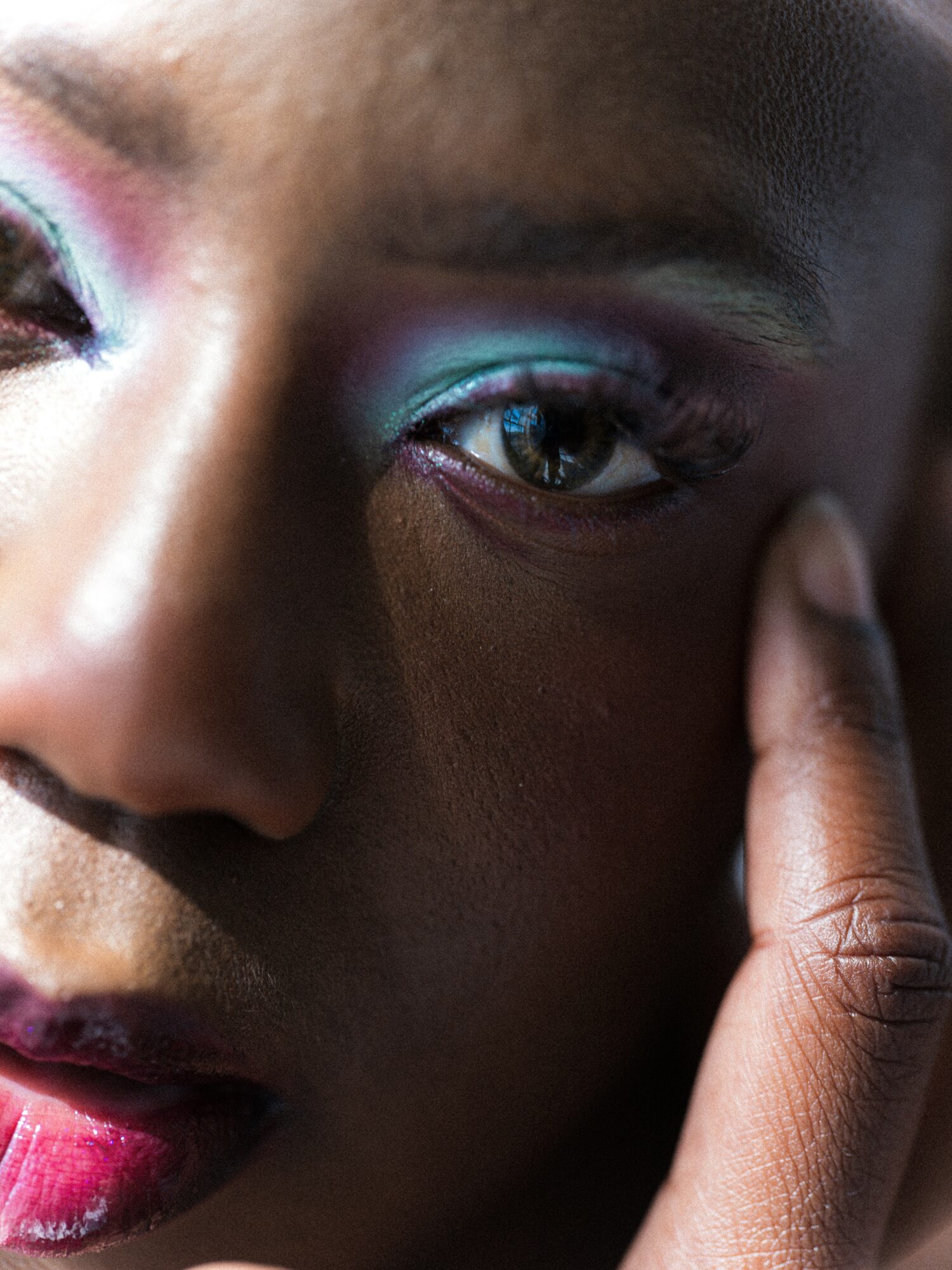 Close-up of a person's face with colorful eye makeup, touching their temple, with visible eye and lips.