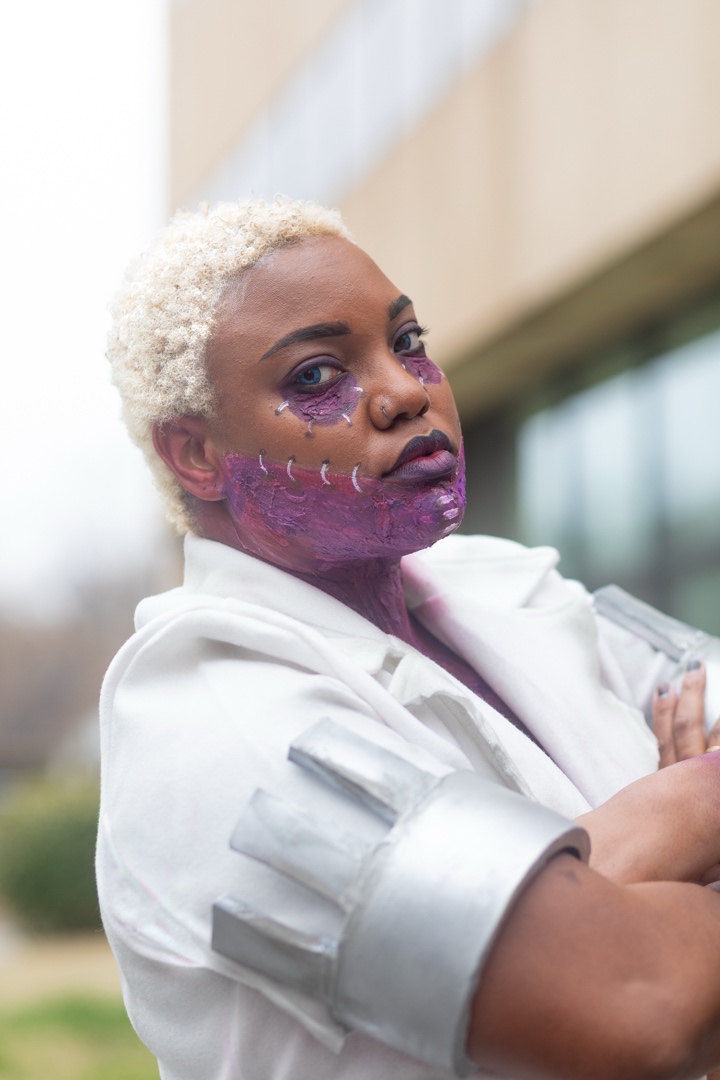 Person with short, curly blonde hair and purple makeup, wearing a white outfit with metallic details, outdoors.