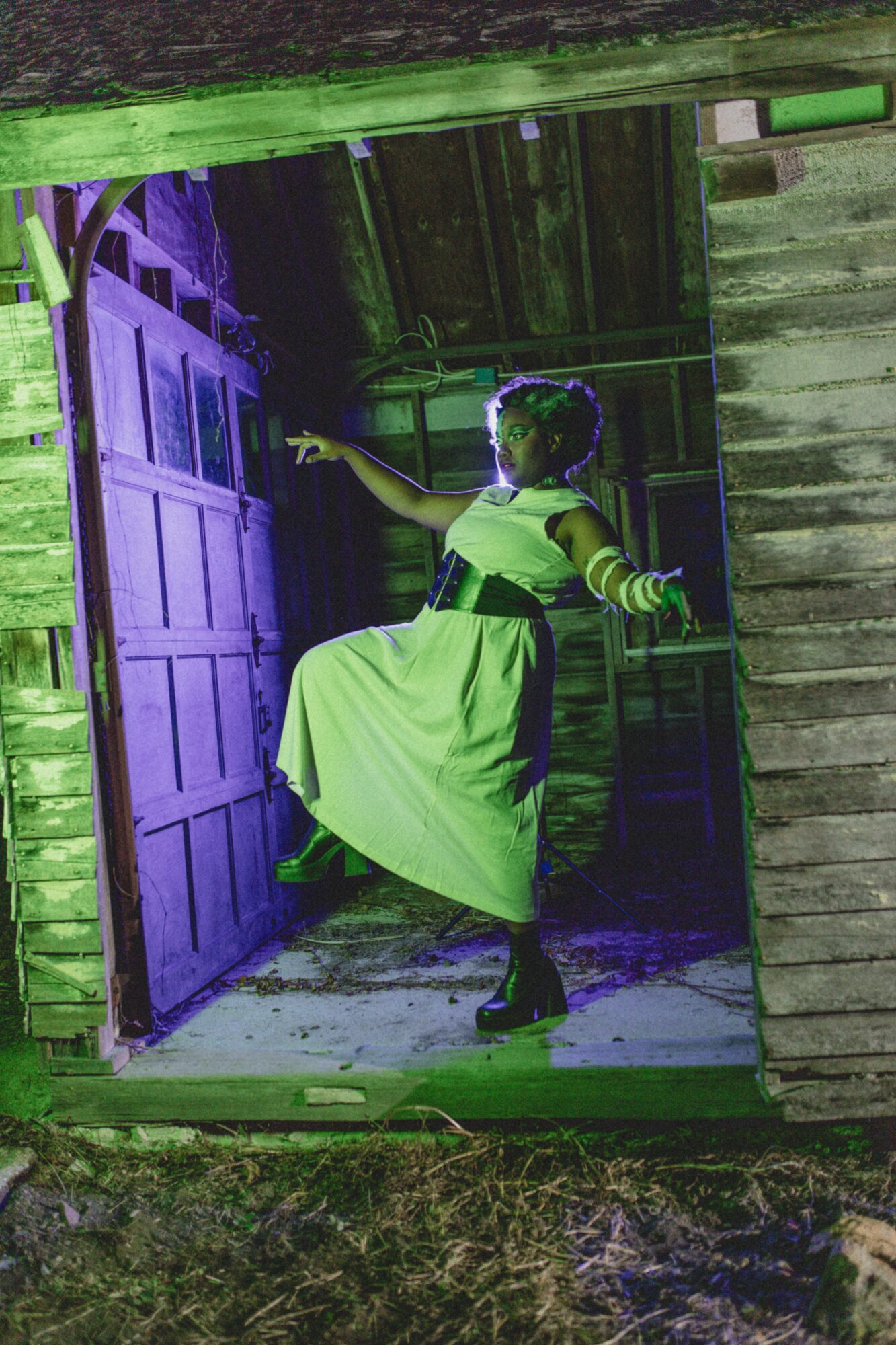 Person in a long dress and headpiece stands in doorway, holding a stick, with purple lighting inside a rustic wooden structure.