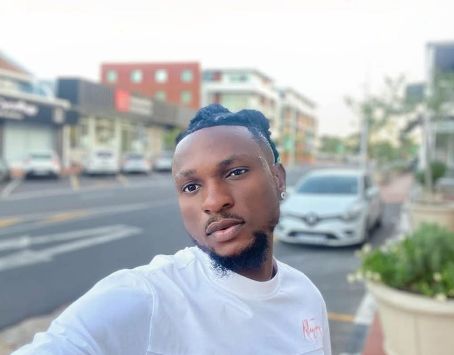 Young man with braided hair and beard taking a selfie outdoors on a city street.
